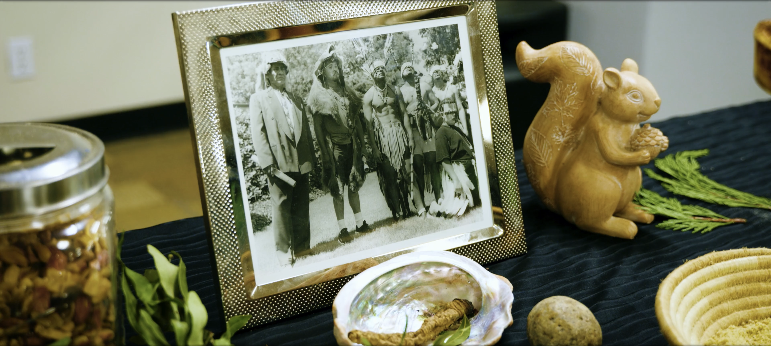 A framed black-and-white photograph of Indigenous people in traditional clothing on a table, surrounded by various natural and cultural artifacts, including a carved rabbit figure, shells, greenery, a jar of mixed nuts, and yellow coiled beads.