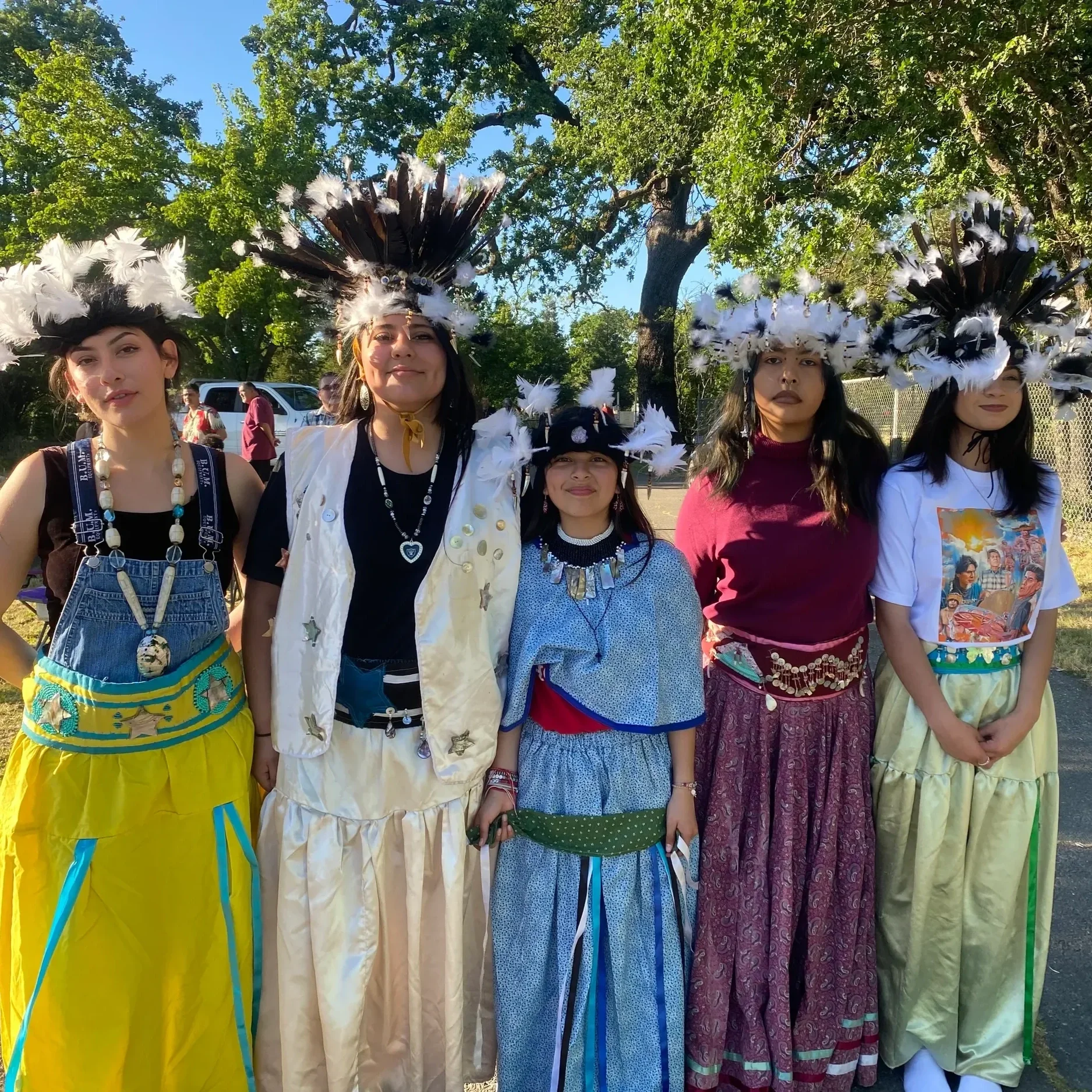 A group of women and children wearing traditional Native American attire with feathered headdresses during an outdoor event in the daytime at a Sonoma County park.