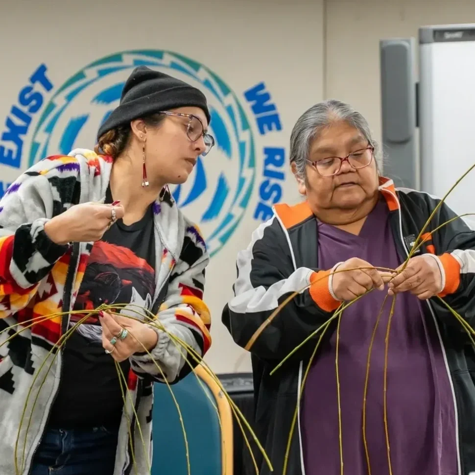 Two women weave reeds for a nonprofit traditional Pomo basket-making workshop in a room with a blue mural in the background.