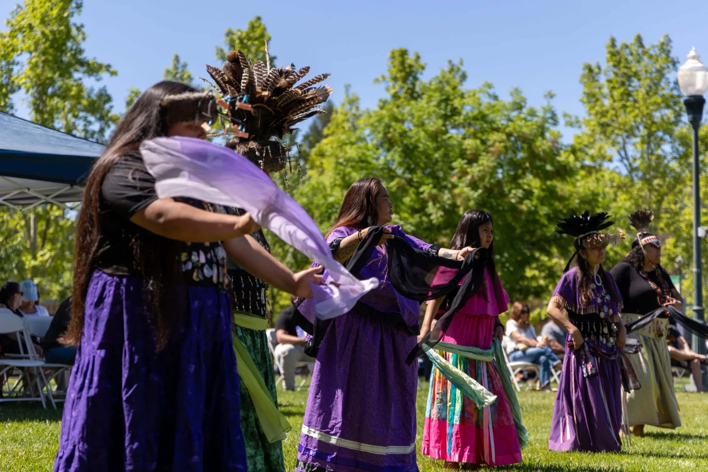 Native women dressed in traditional clothing perform a dance outdoors under a clear blue sky, with green trees and an audience in the background at a Native nonprofit event in Sonoma County.