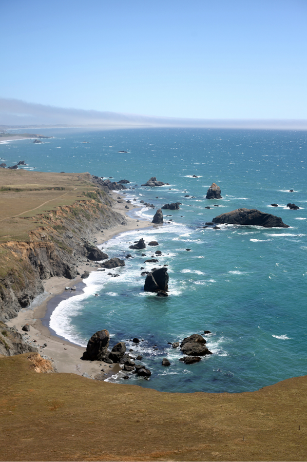 Sonoma coastline with rocky shoreline, waves, green cliffs, and a clear blue sky.