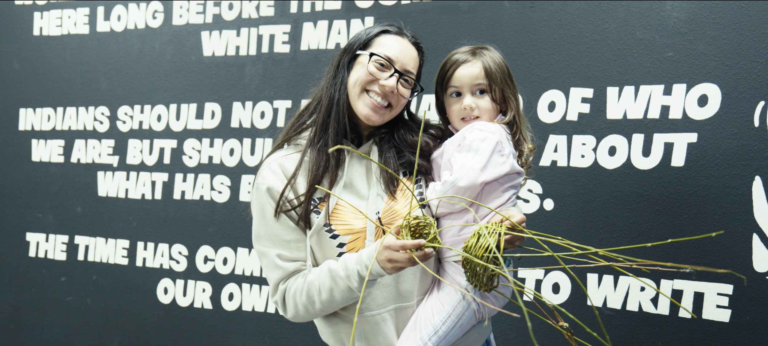 A Native woman with glasses and a young girl stand in front of a black wall with white text. The woman is smiling and holding a handmade basket while the girl is looking at the camera with a neutral expression.