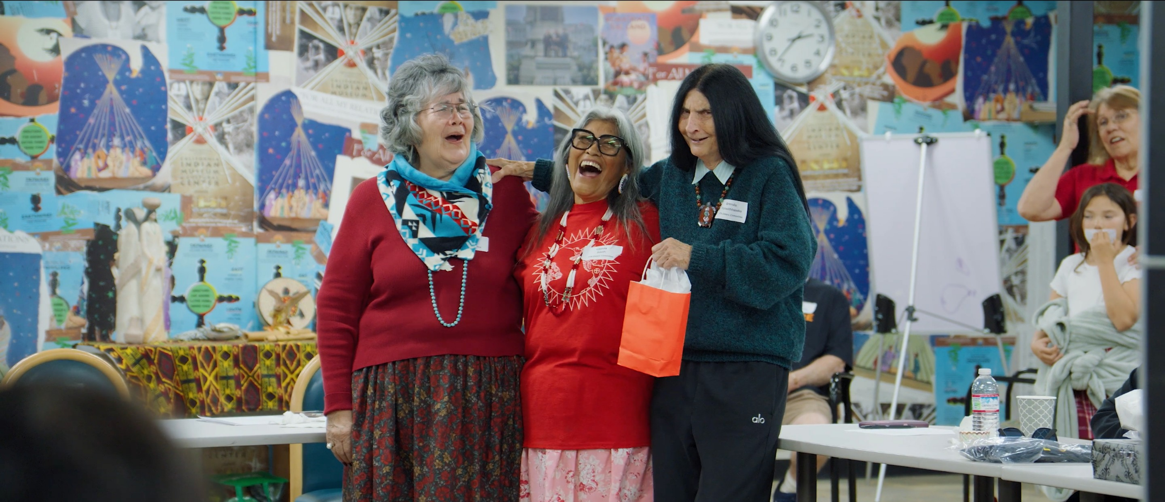 Three native elder women smile at an indoor elders' gathering, surrounded by colorful posters and artwork on the walls.