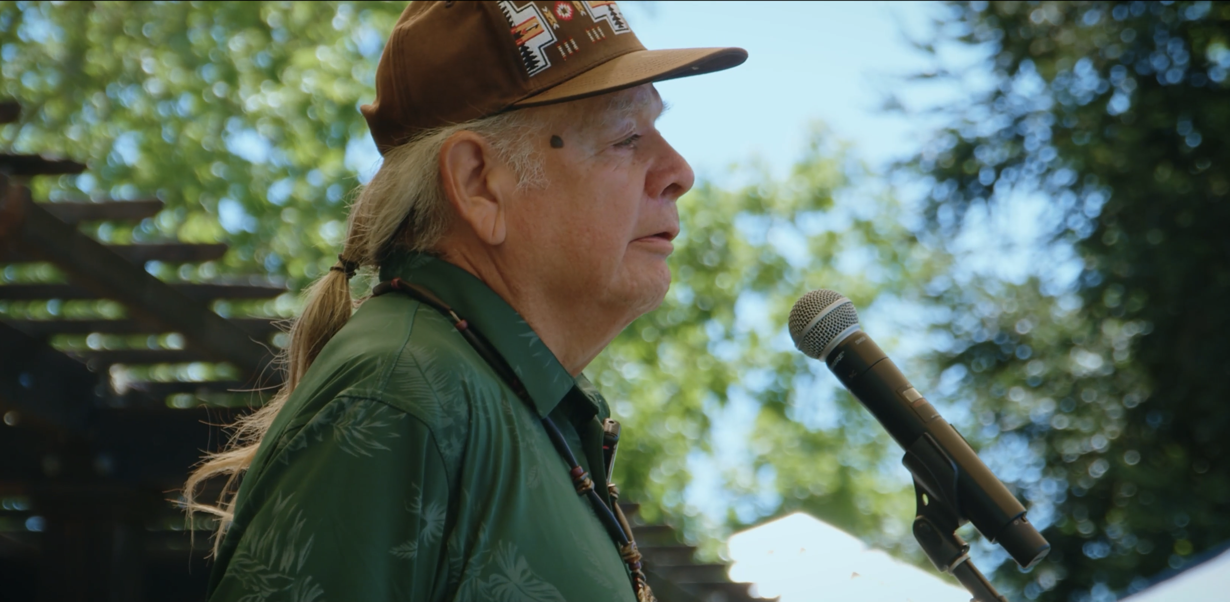 An older Native man with long gray hair in a ponytail, wearing a brown hat and a green patterned shirt, speaking into a microphone outdoors during daytime with green trees and a wooden structure in the background.