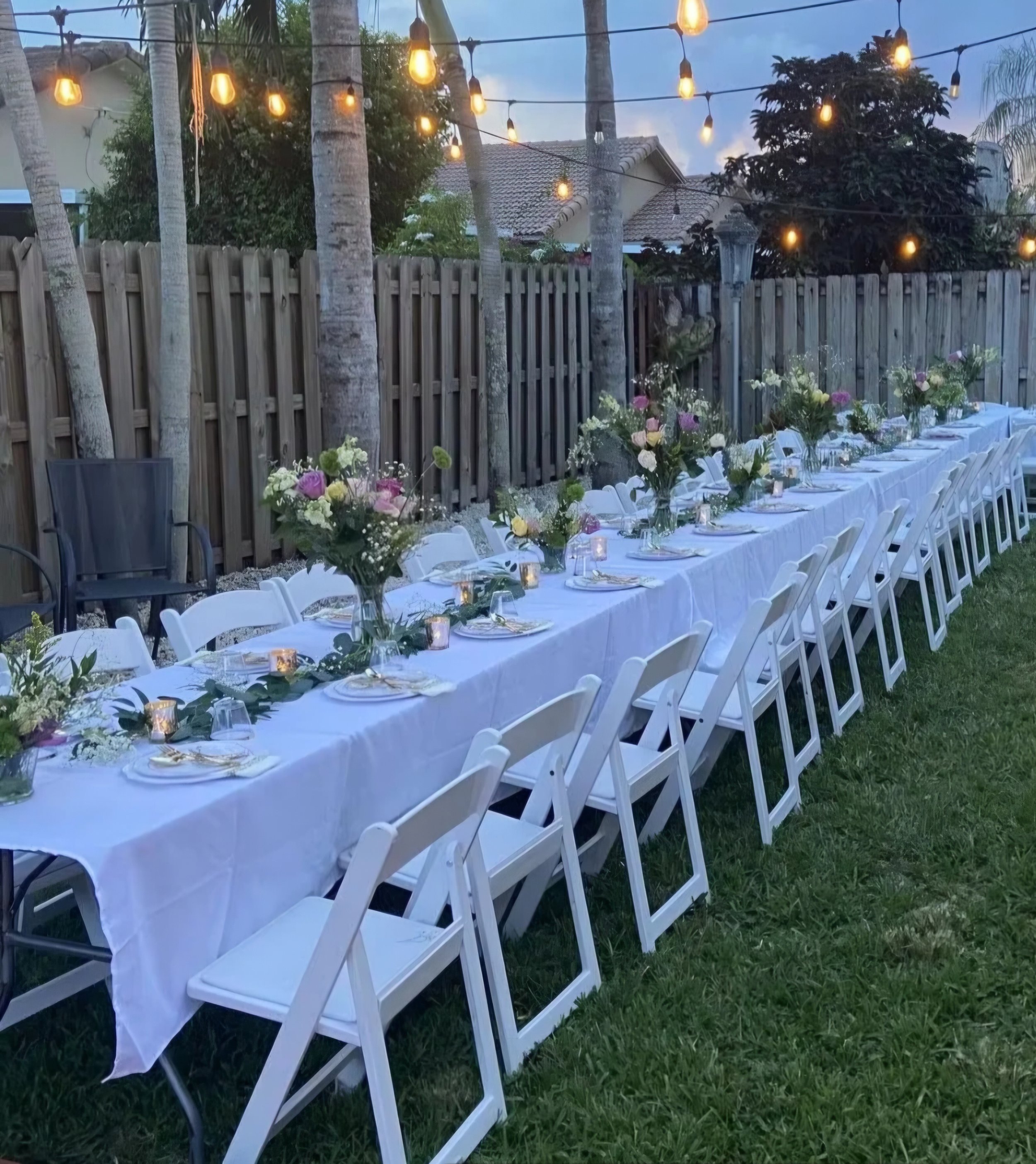 Outdoor dinner setup with a long table covered in white tablecloth, decorated with floral arrangements and candles, set against a backyard fence with hanging string lights and trees.