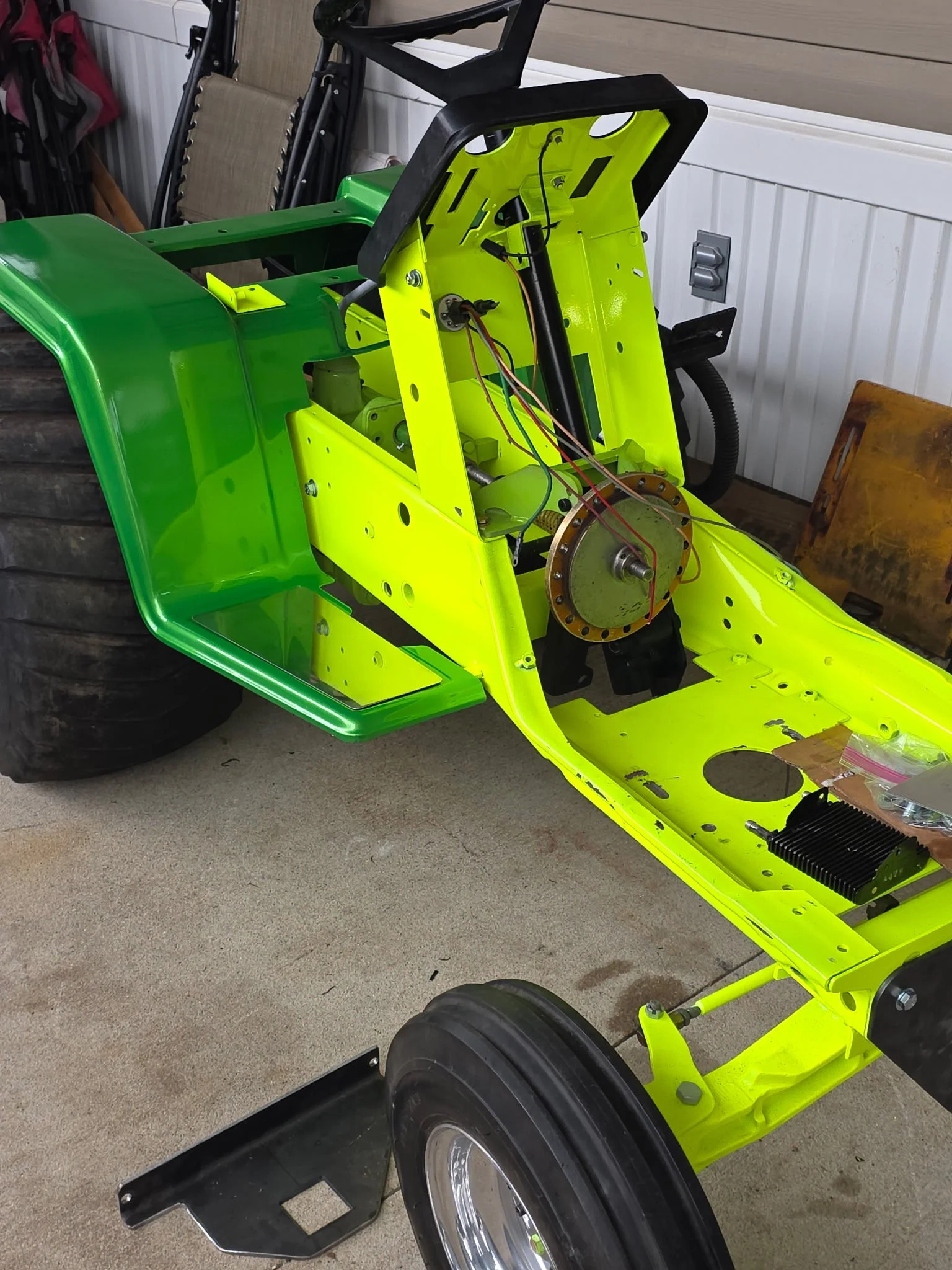 A partially assembled garden tractor with a bright yellow frame, black tires, and exposed wiring inside a workshop or garage. There's a large tire in the background, and various tools and materials nearby.