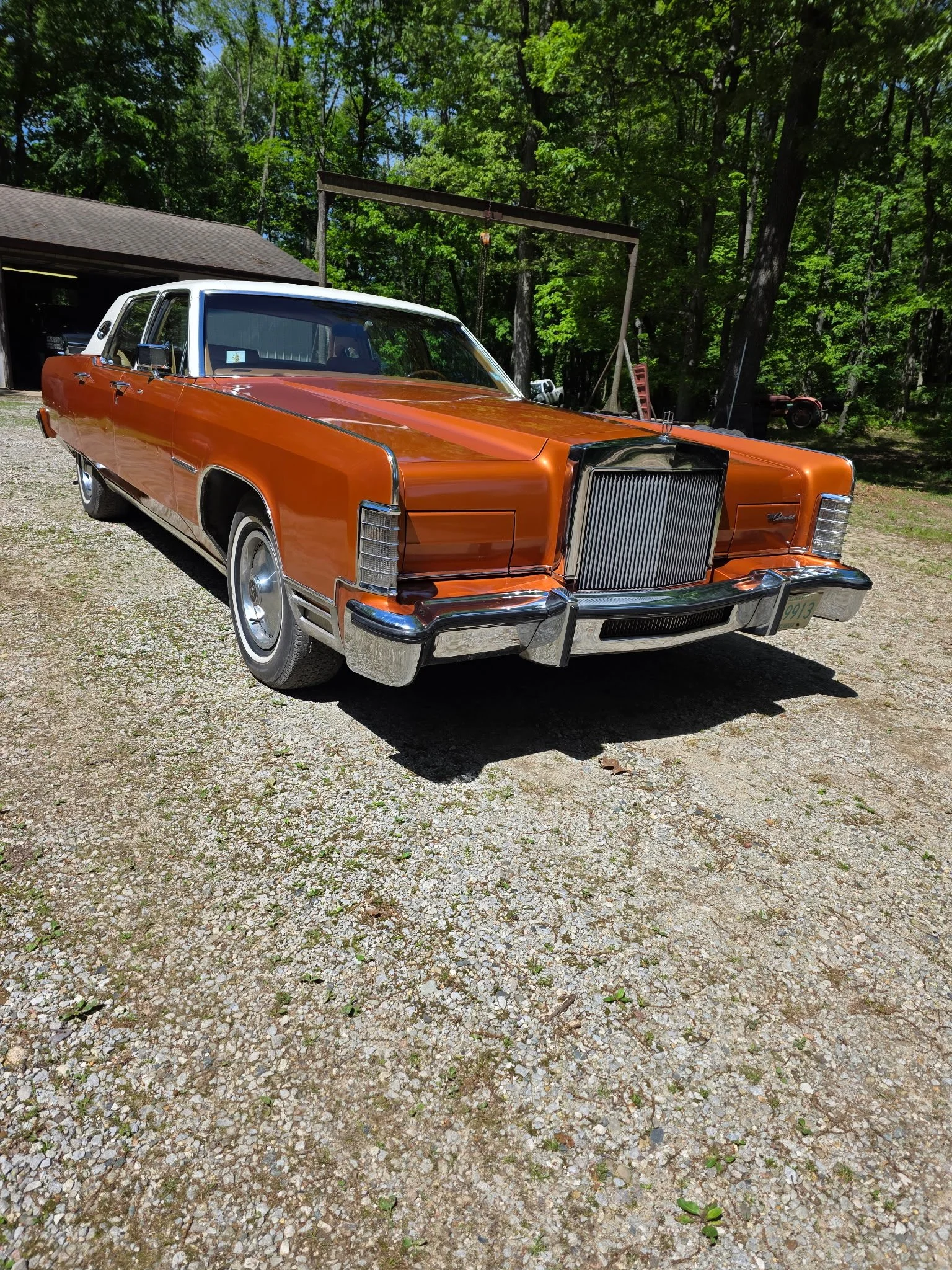 An orange vintage car with a white roof parked on a gravel surface in a wooded area, with trees and a garage in the background.