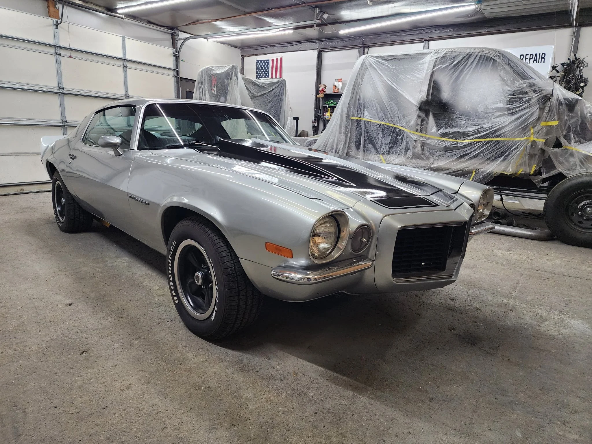A silver Classic Chevrolet Camaro parked inside a garage next to a covered vehicle and automotive tools on shelves, with an American flag hanging on the wall.