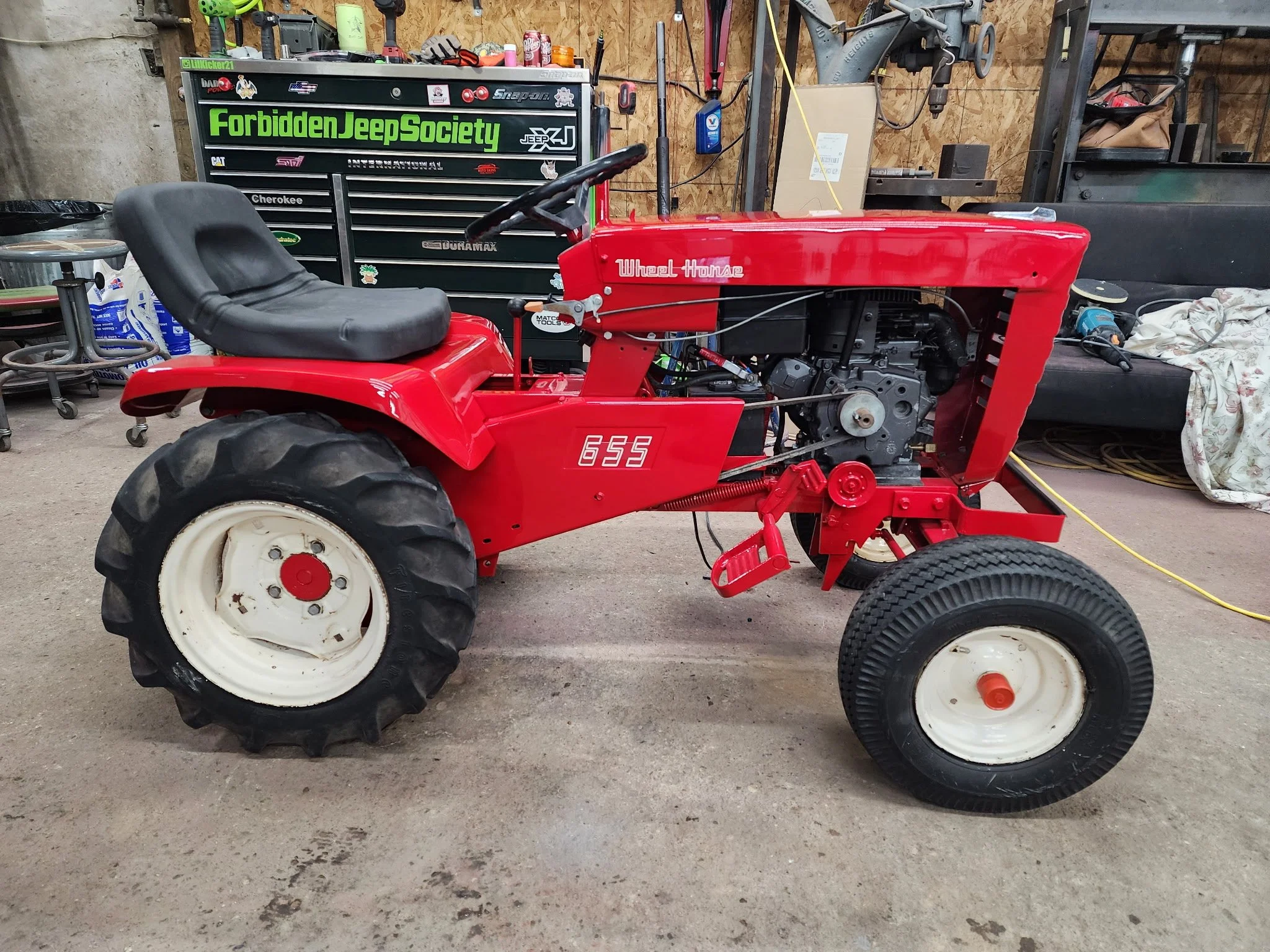 A red tractor in a garage or workshop with a black seat, large rear tires, and a visible engine. There are tools, a black tool chest with stickers, a workbench, and various equipment in the background.