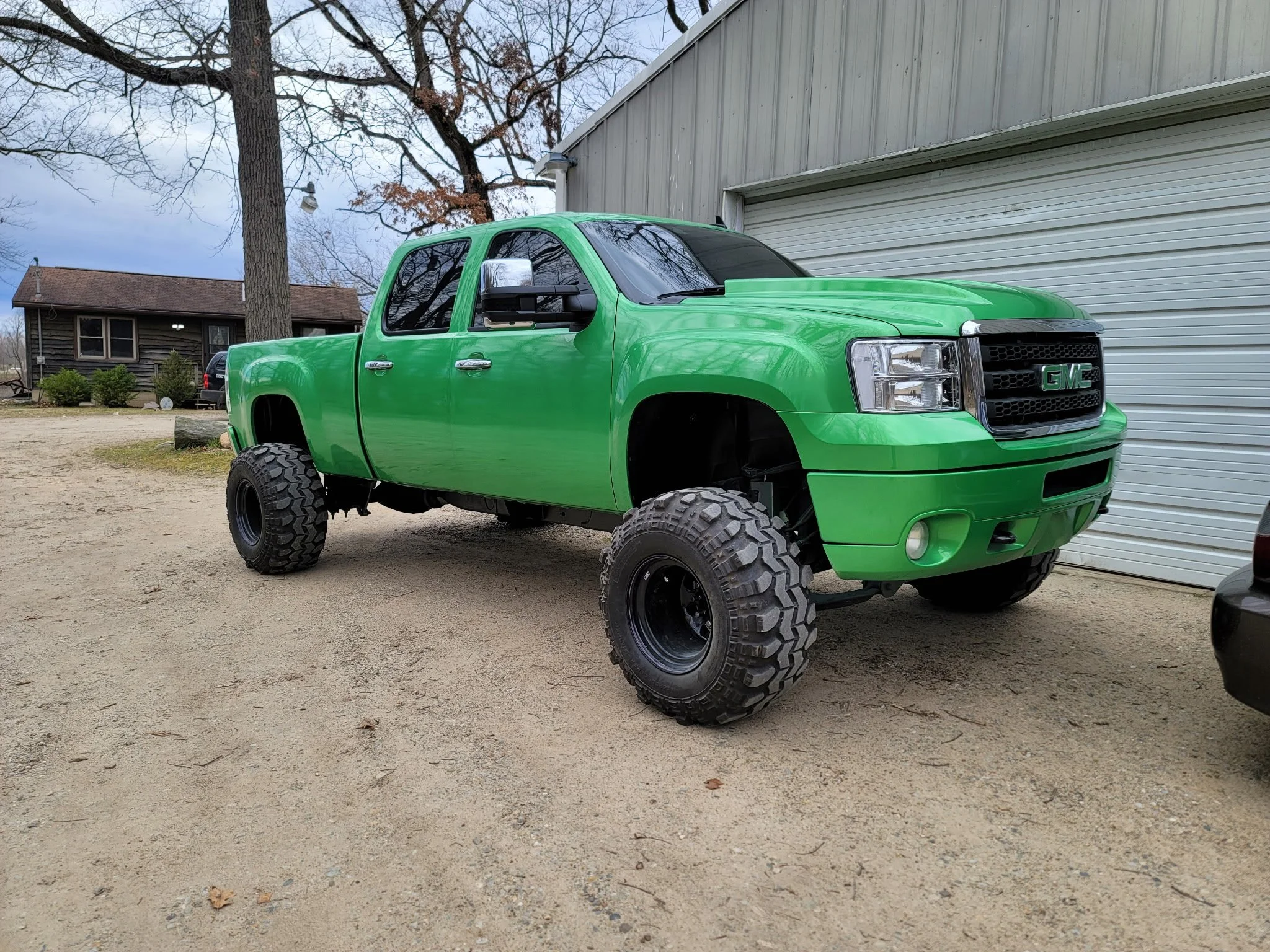 A bright green lifted GMC pickup truck with large off-road tires parked in a rural driveway beside a house with a metal garage door. The background shows a leafless tree, a house, and a cloudy sky.