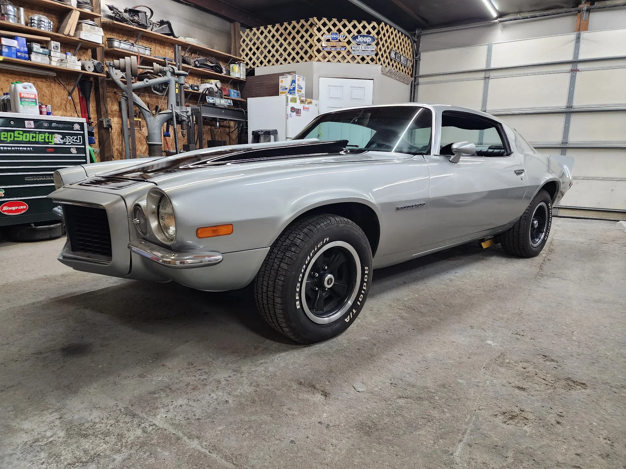 A silver vintage muscle car parked inside a garage with wood-paneled walls, shelves filled with car parts and tools, and various automotive stickers and signs on the walls.