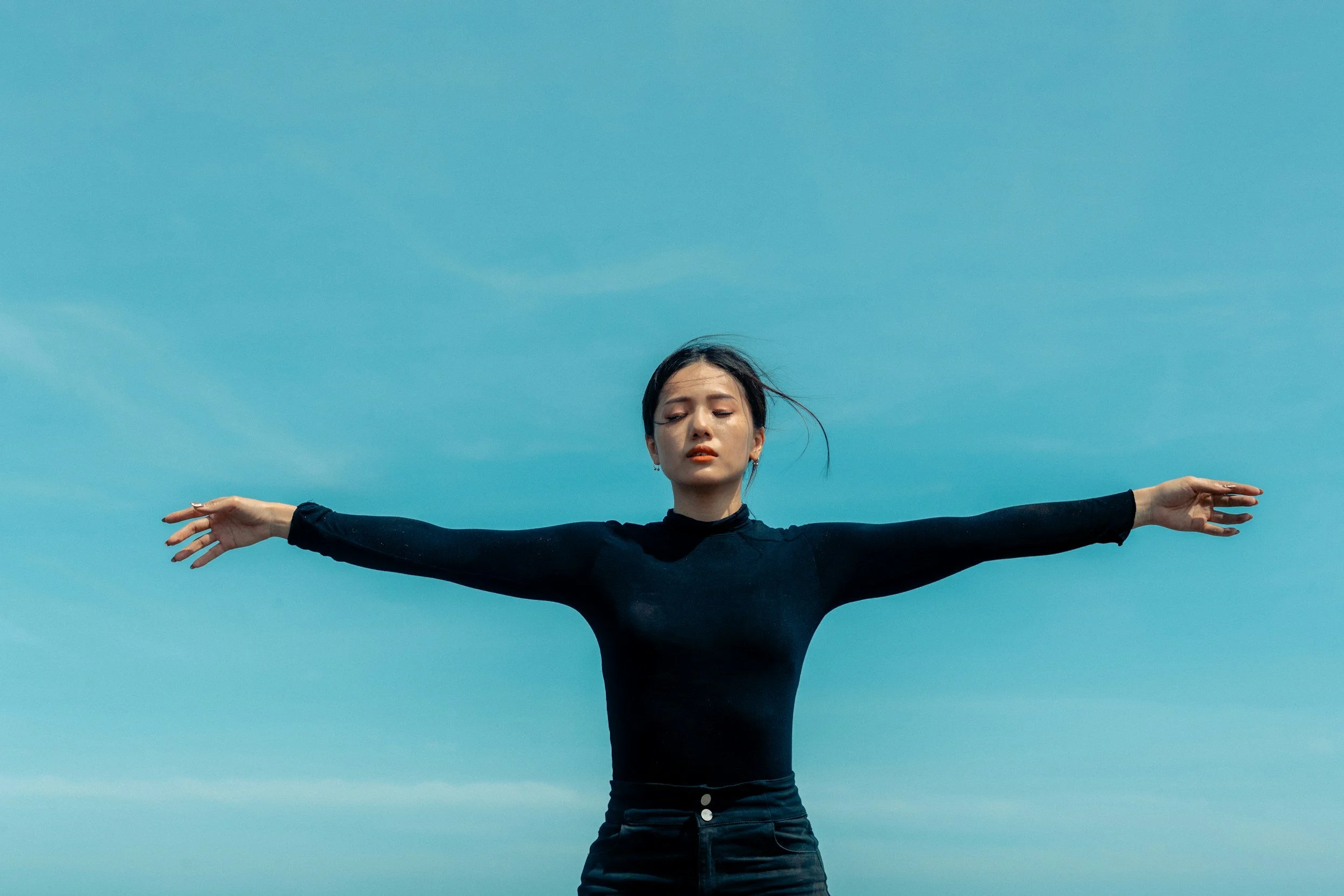 A woman with dark hair, wearing a black long-sleeve top, has arms outstretched against a clear blue sky, looking peaceful.