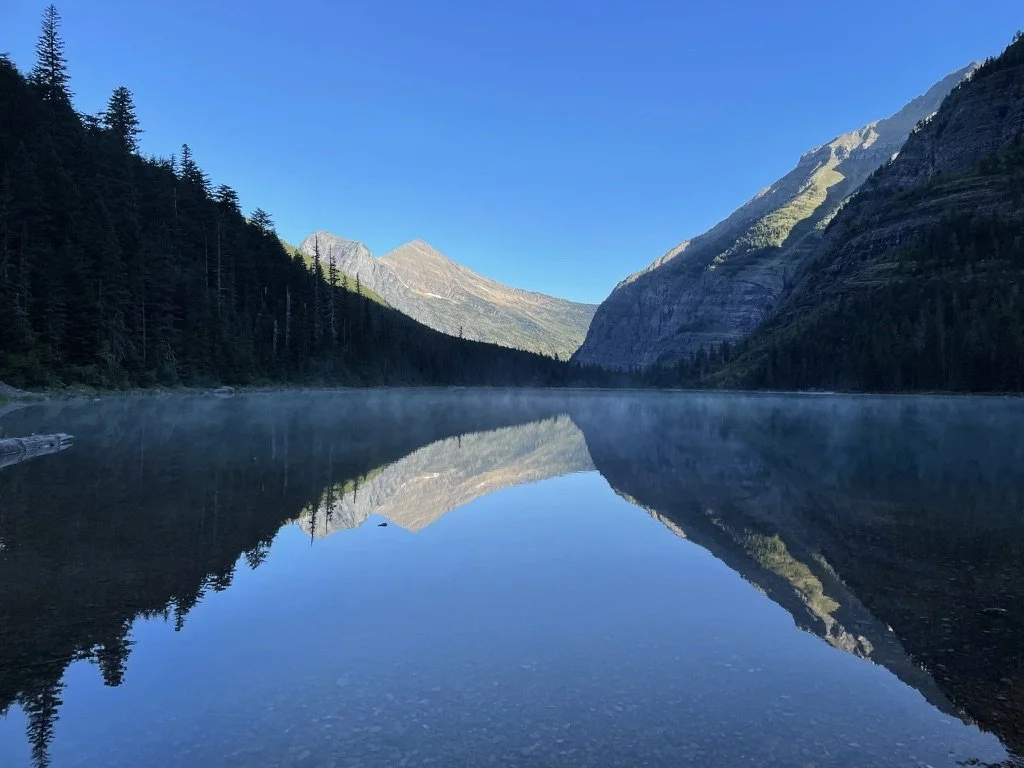 A calm mountain lake reflecting the surrounding forested mountains and clear blue sky.