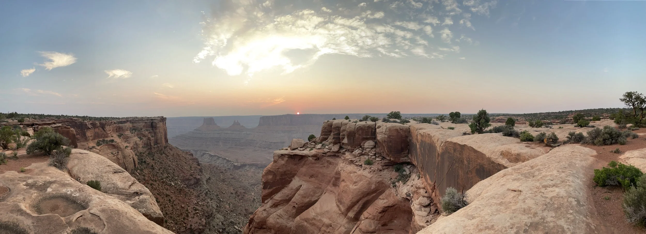 A panoramic view of Grand Canyon at sunset, showing layered red rock formations, cliffs, and sparse desert vegetation under a partly cloudy sky.