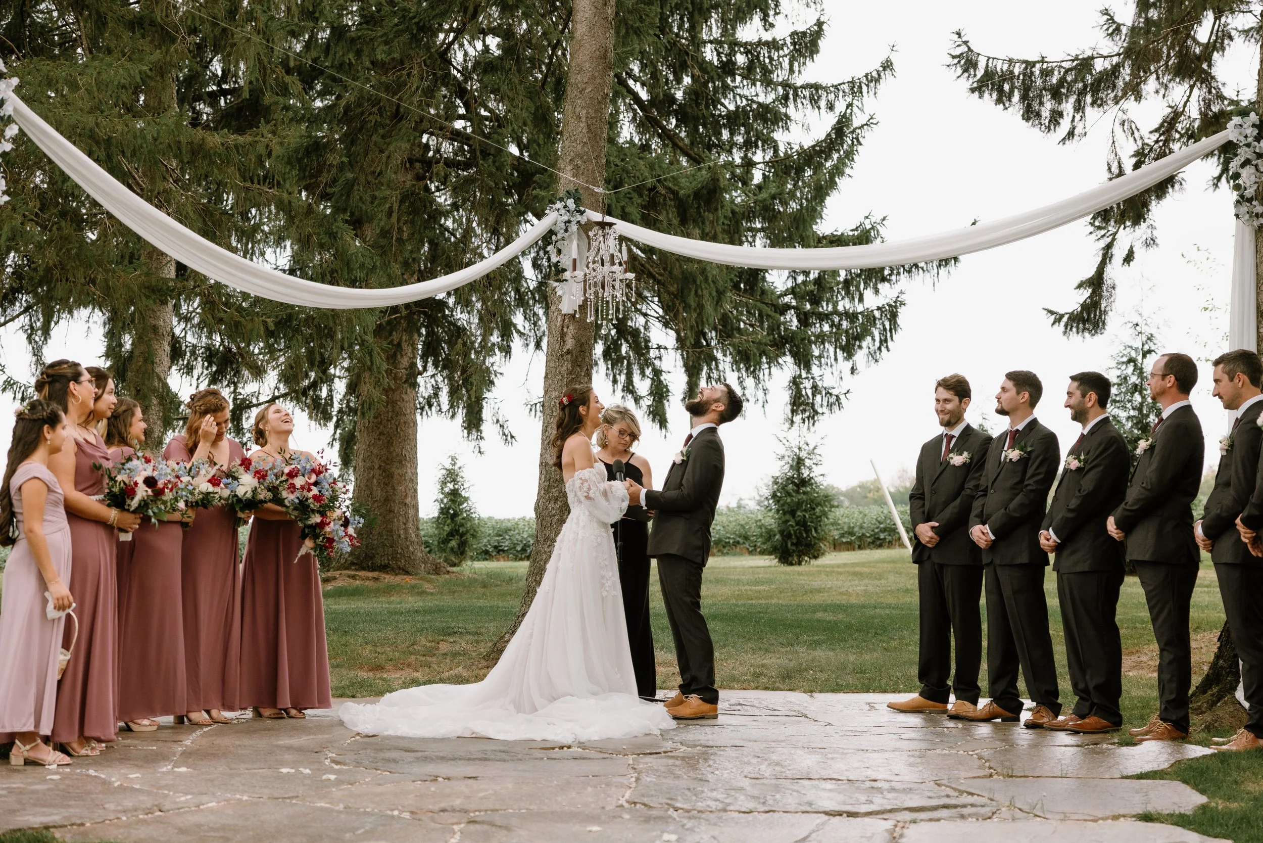 A wedding ceremony outdoors with a bride and groom exchanging vows, surrounded by bridesmaids in pink dresses holding bouquets and groomsmen in suits. The setting features trees and a decorative draped fabric with a chandelier hanging from above.