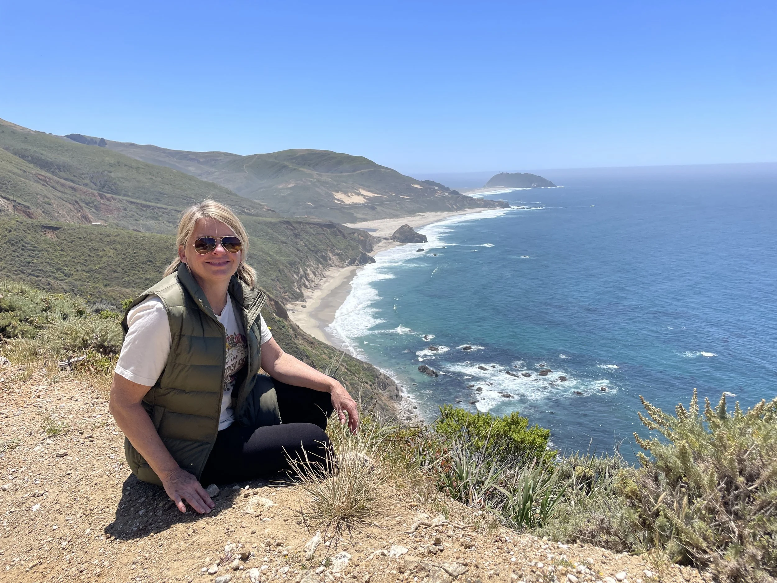 A woman in sunglasses and a puffy vest sitting on a dirt path overlooking the ocean, with green hills and rocky coast in the background.