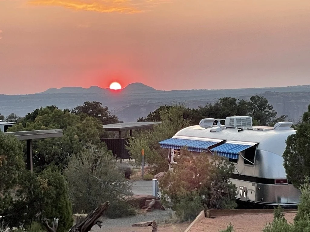 A scenic sunset over mountains, with an RV parked among trees and bushes in the foreground.