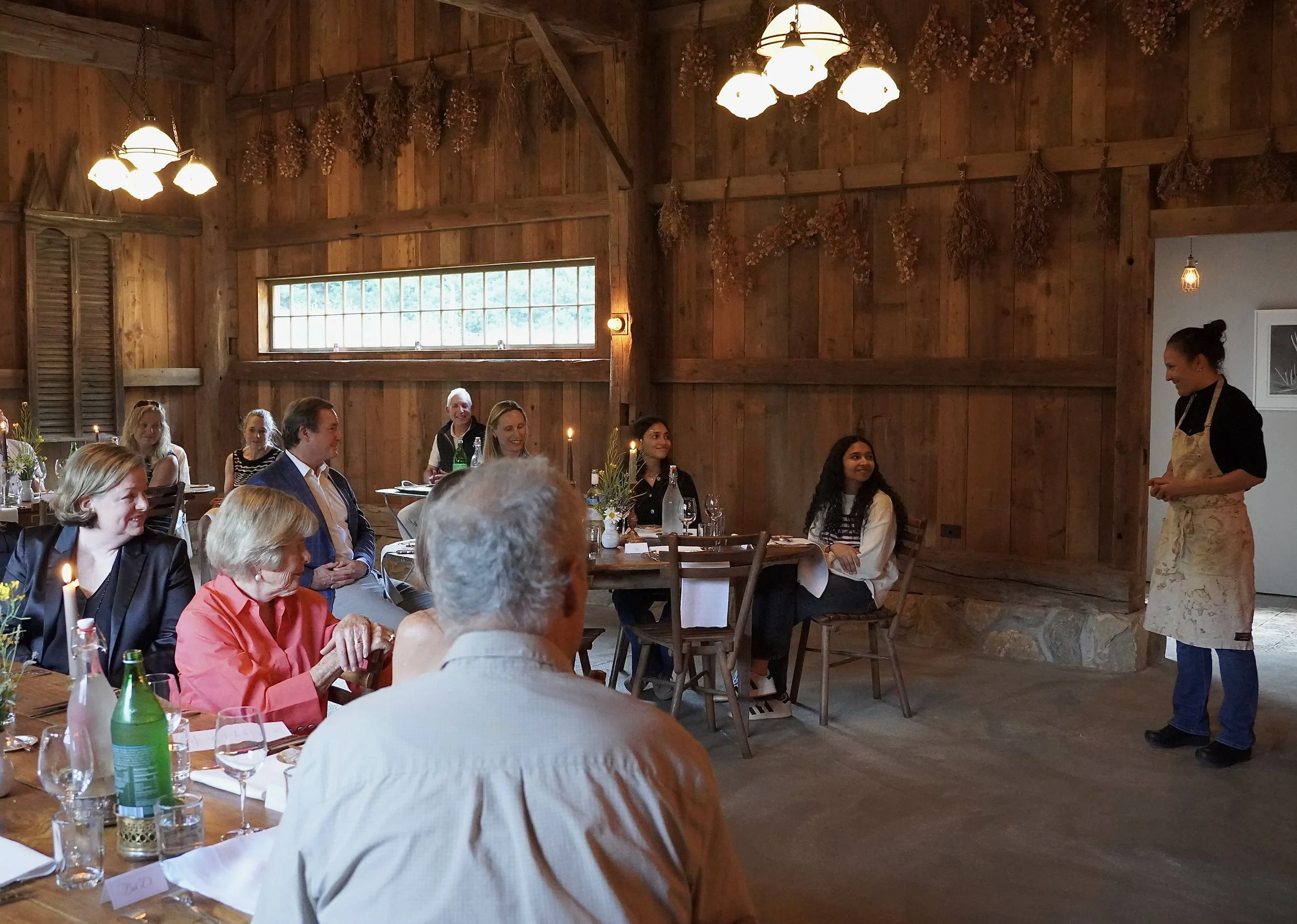 A group of people are gathered indoors in a rustic wooden room, attending a presentation or speech. The room is decorated with dried flowers hanging from the ceiling, and warm lighting from chandeliers and small lamps. The audience is seated at tables with drinks and candles, and a woman stands to the right, speaking to the group.
