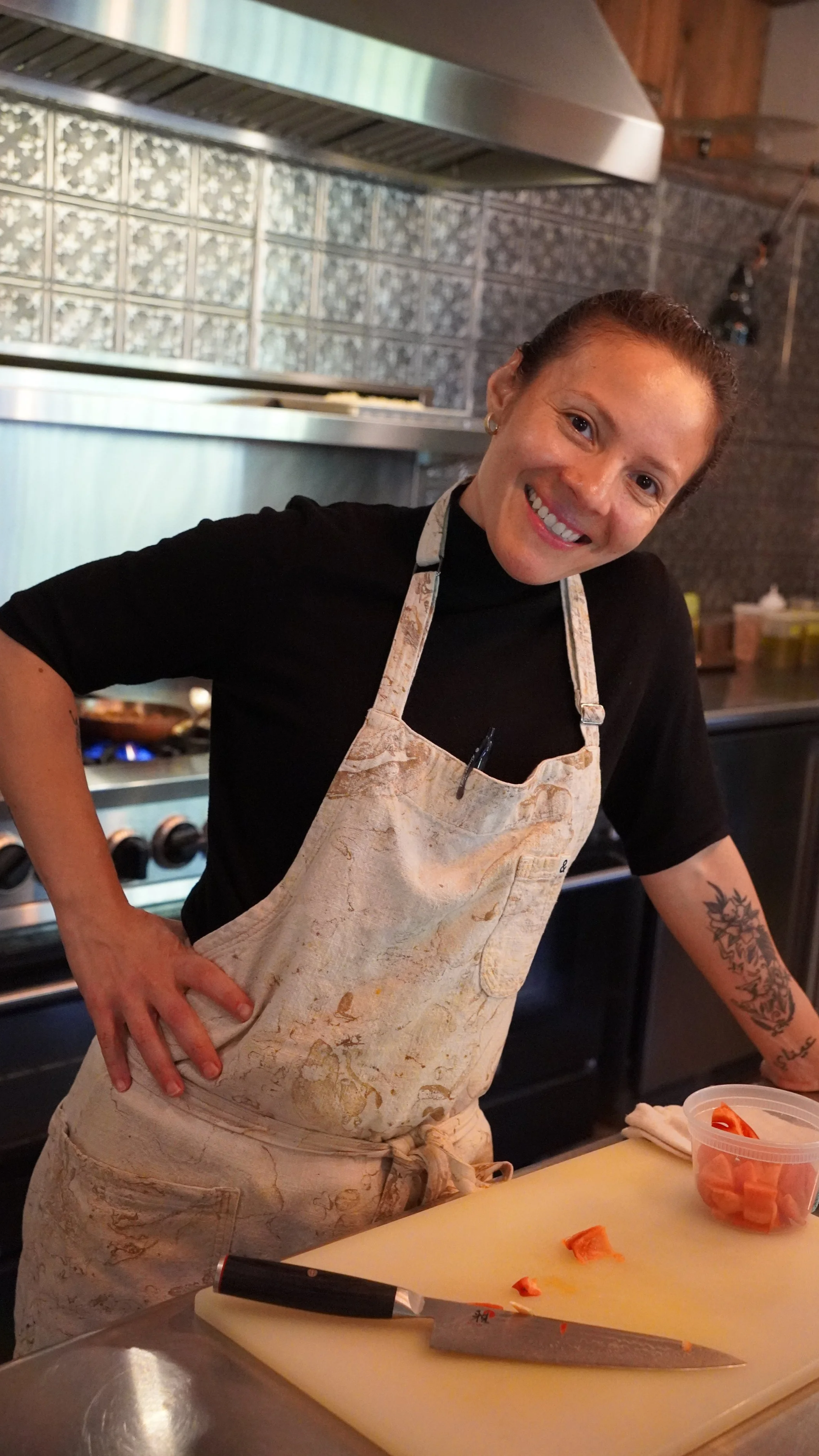 A woman wearing a black shirt and a beige apron, smiling and standing in a kitchen with metallic and tiled backgrounds. She is preparing a dish with chopped vegetables on a yellow cutting board, with a knife and a small container of chopped vegetables nearby.