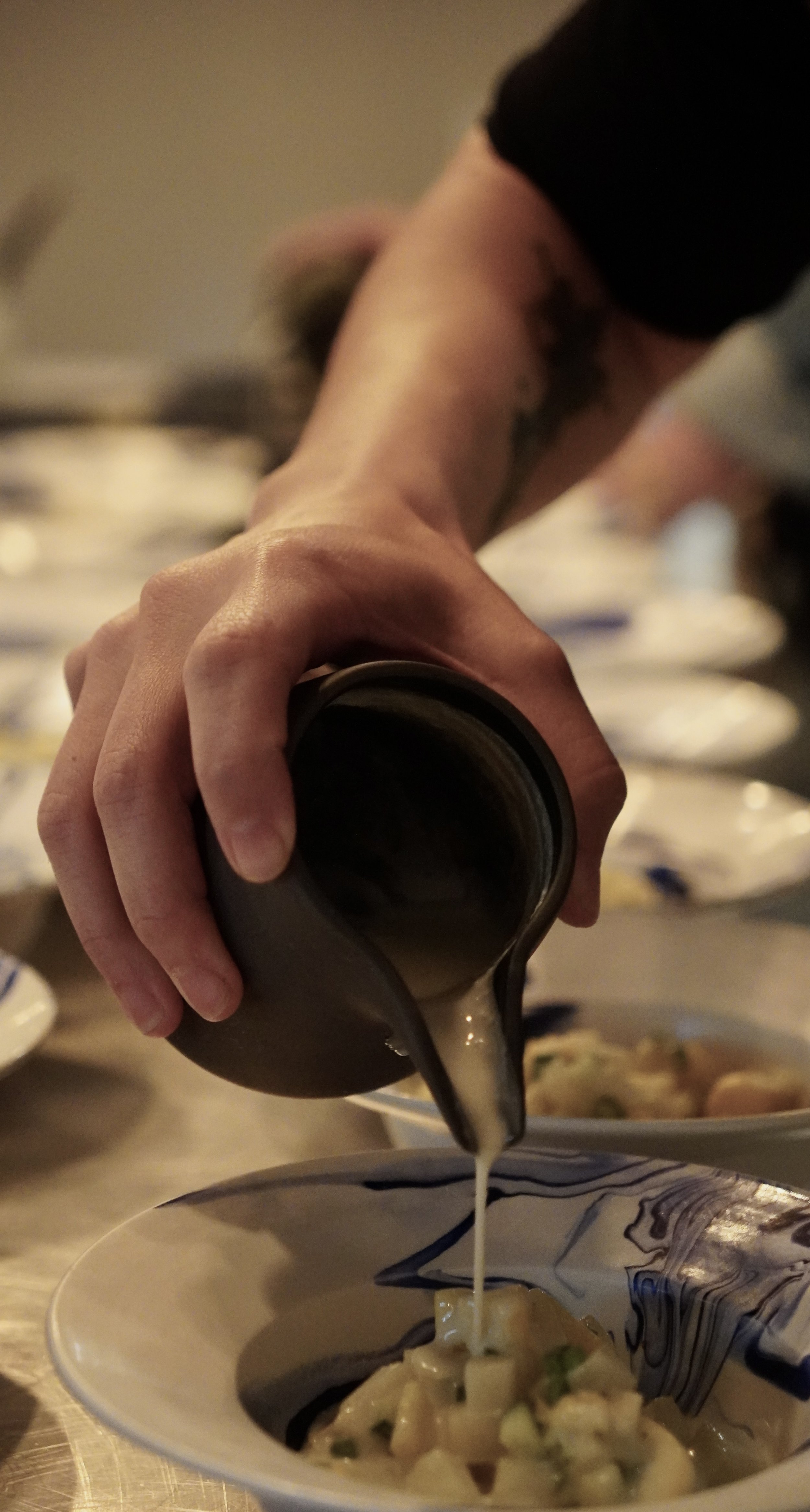 A person pouring cream or sauce into a bowl of pasta with vegetables.