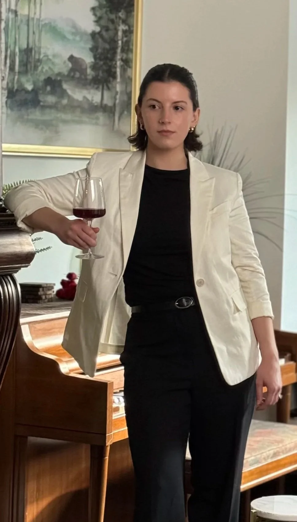 A woman with dark hair wearing a white blazer and black top holds a glass of red wine in a living room or dining area.