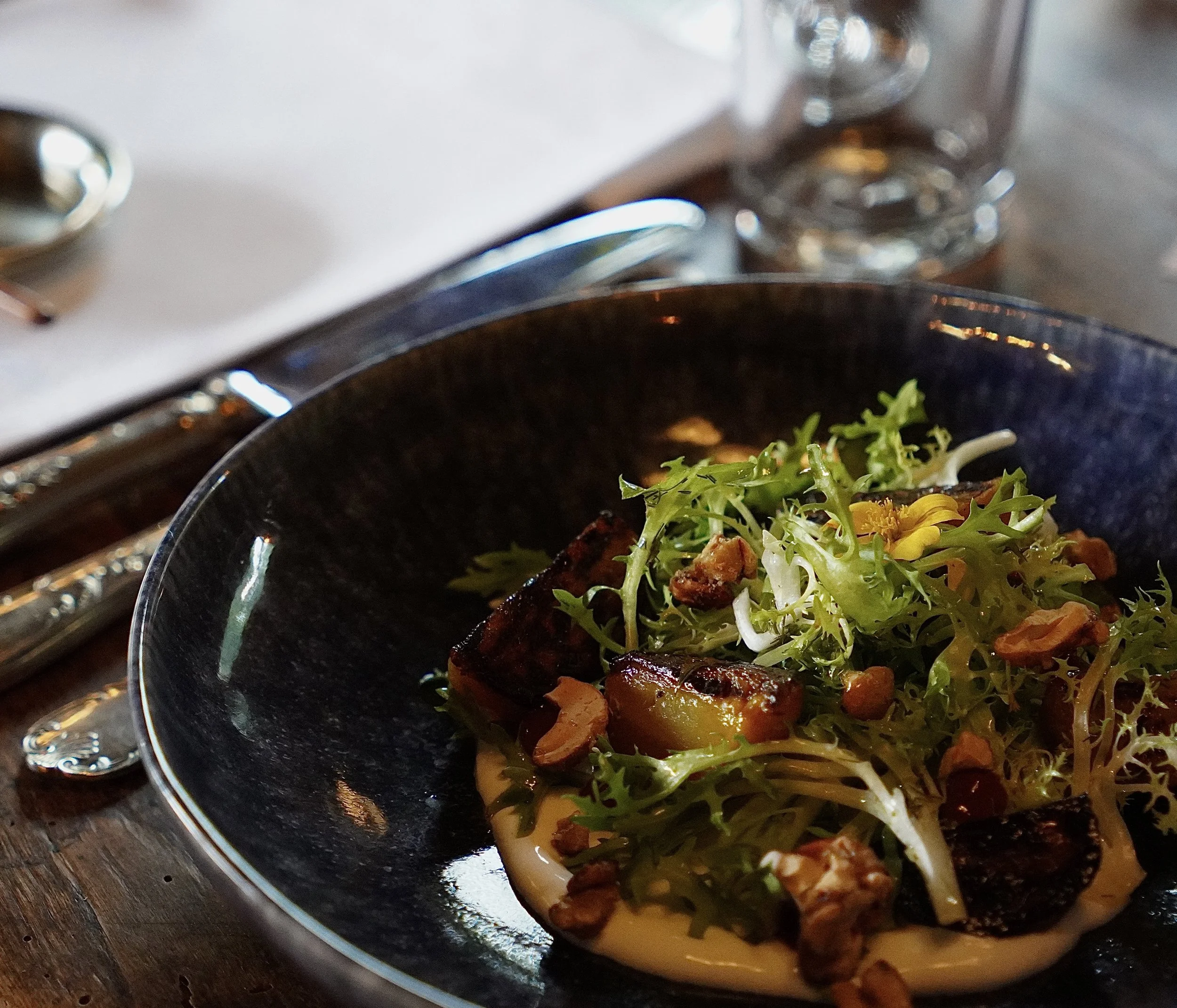 Close-up of a bowl of salad with greens, what appears to be grilled or roasted vegetables, and possibly pieces of meat, on a dark plate. Part of a silver utensil is visible next to the bowl, on a wooden surface.