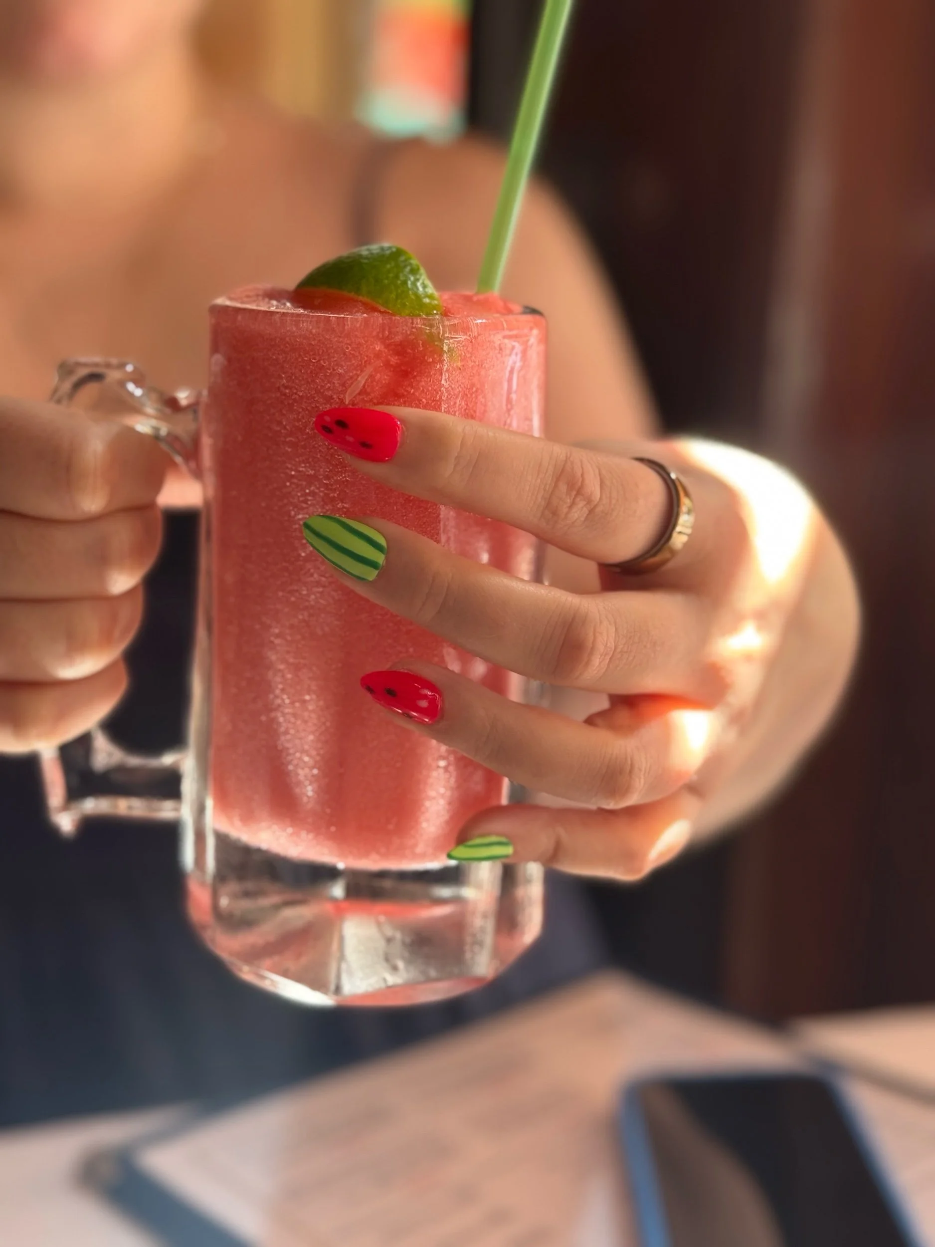 Close-up of a person's hand holding a pink frosted drink garnished with lime, with bright watermelon press-on nails.