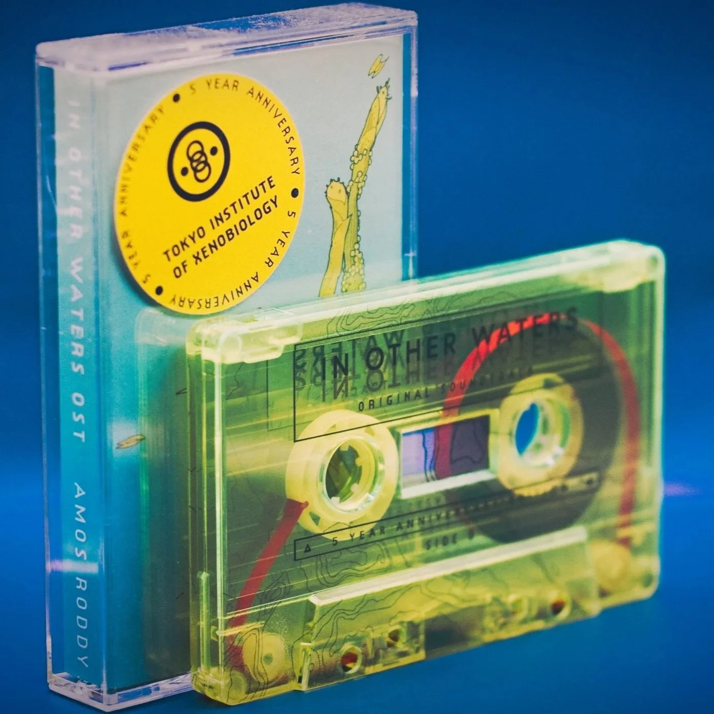A cassette tape of the In Other Waters soundtrack by Amos Roddy and its case, arranged against a blue background.