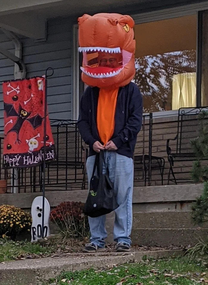 Steven standing in a front yard on Halloween wearing a giant, orange T. Rex head.