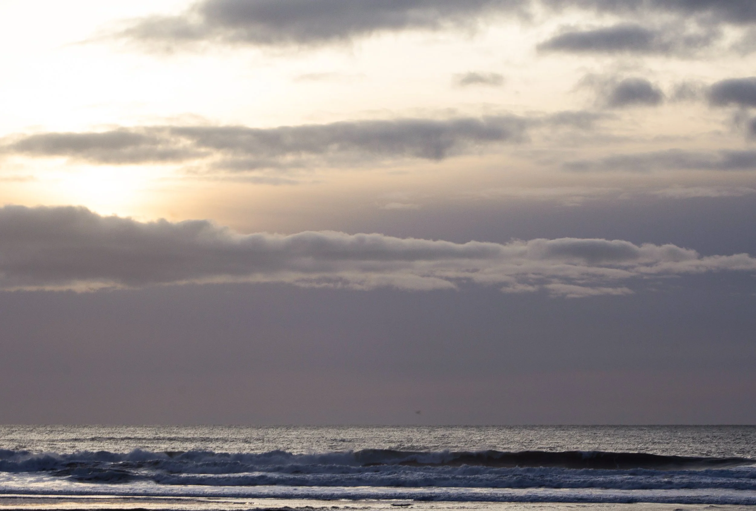 A cloudy beach scene at sunset or sunrise with a dark ocean, waves, and a sky filled with gray clouds.