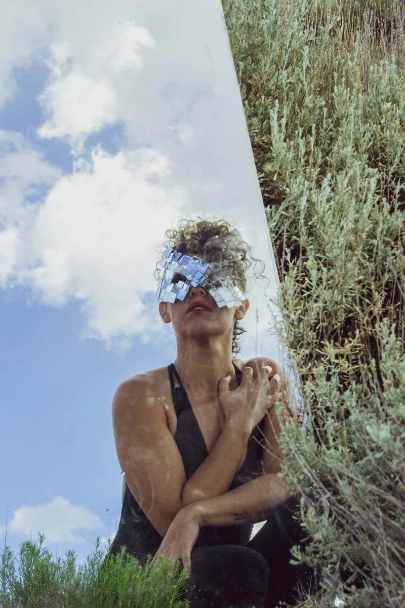 A woman with curly hair wearing a black tank top, looking into a mirror, with her hand on her chest. She has a reflective accessory over her eyes, and the photo captures the sky with clouds behind her and some green plants in front of her.
