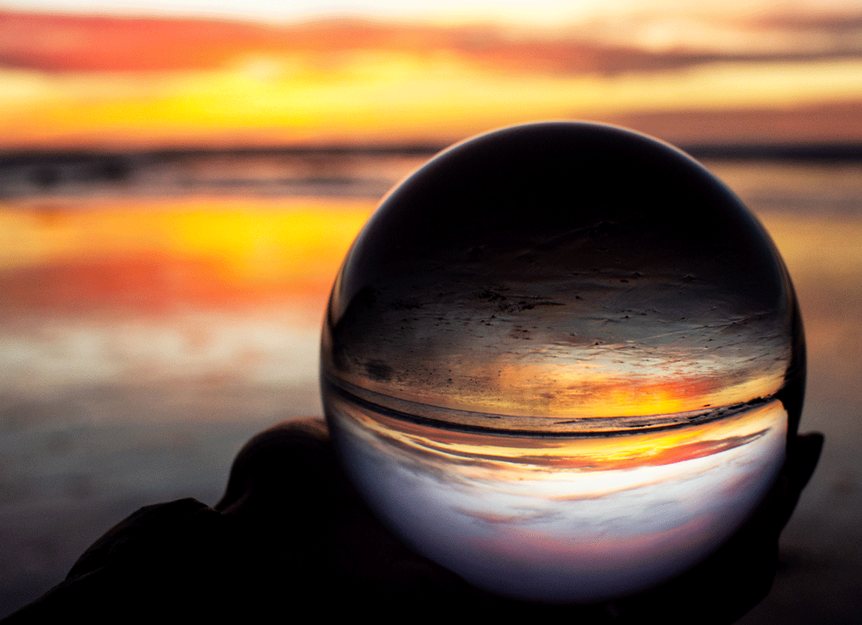 A glass sphere on a beach reflecting a sunset over the ocean with colorful clouds and water.