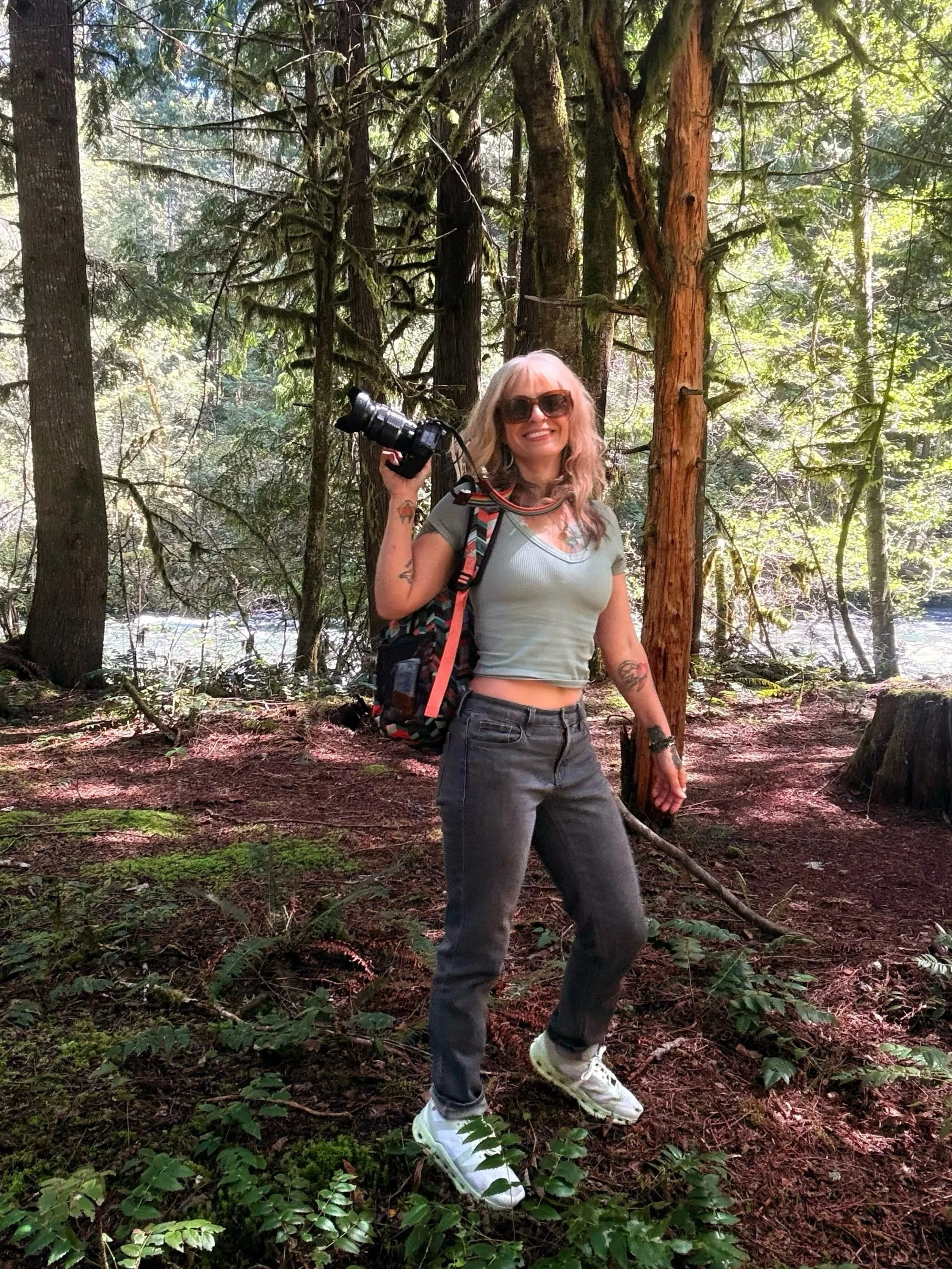 A woman with glasses and blonde hair smiling, holding a camera, standing in a forest with trees and greenery.