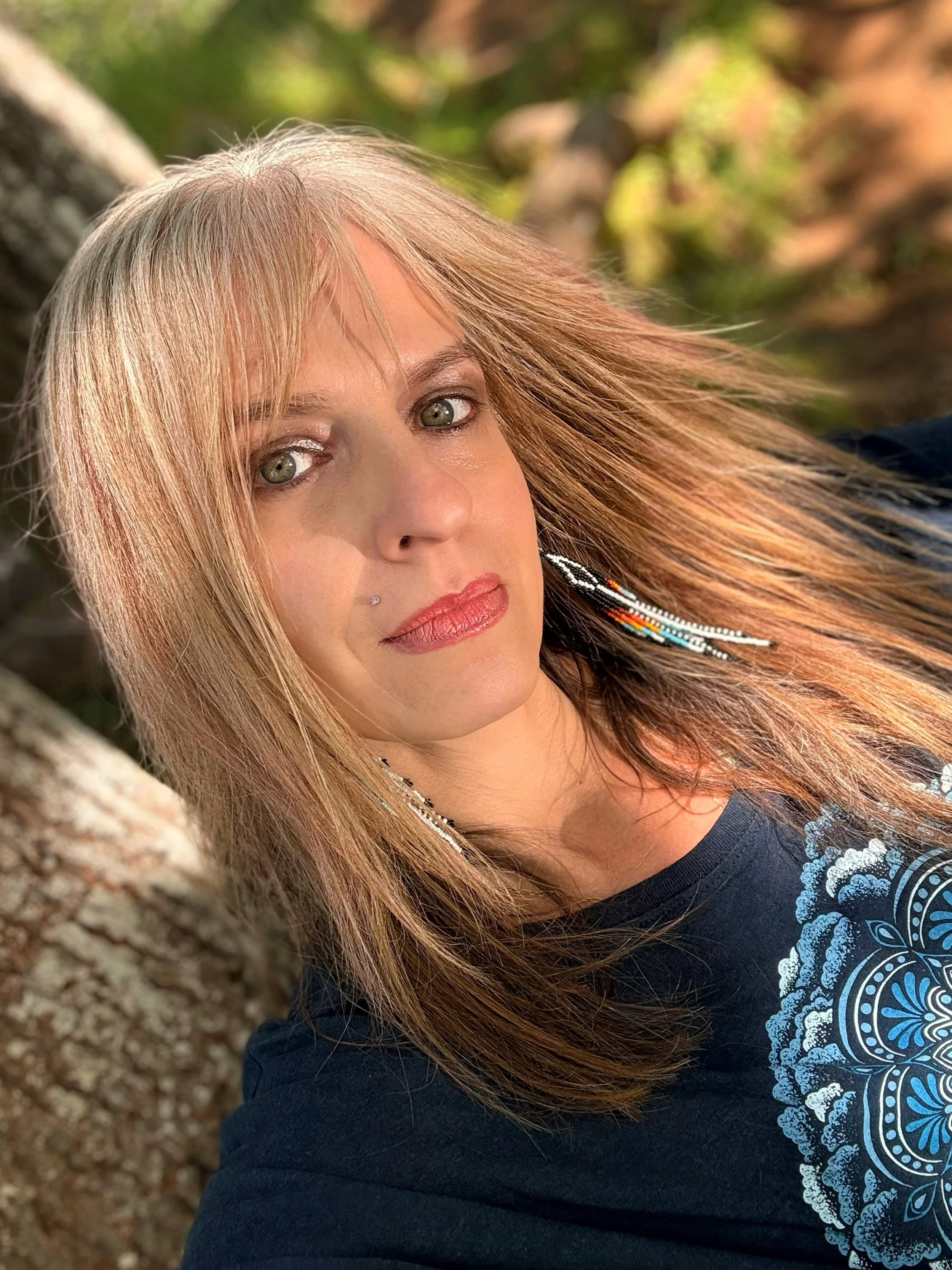 Close-up of a woman with long reddish-blonde hair, light skin, and gray eyes, wearing makeup and a black shirt with a blue and white decorative pattern. She is outdoors near a tree, with blurred green and brown background.