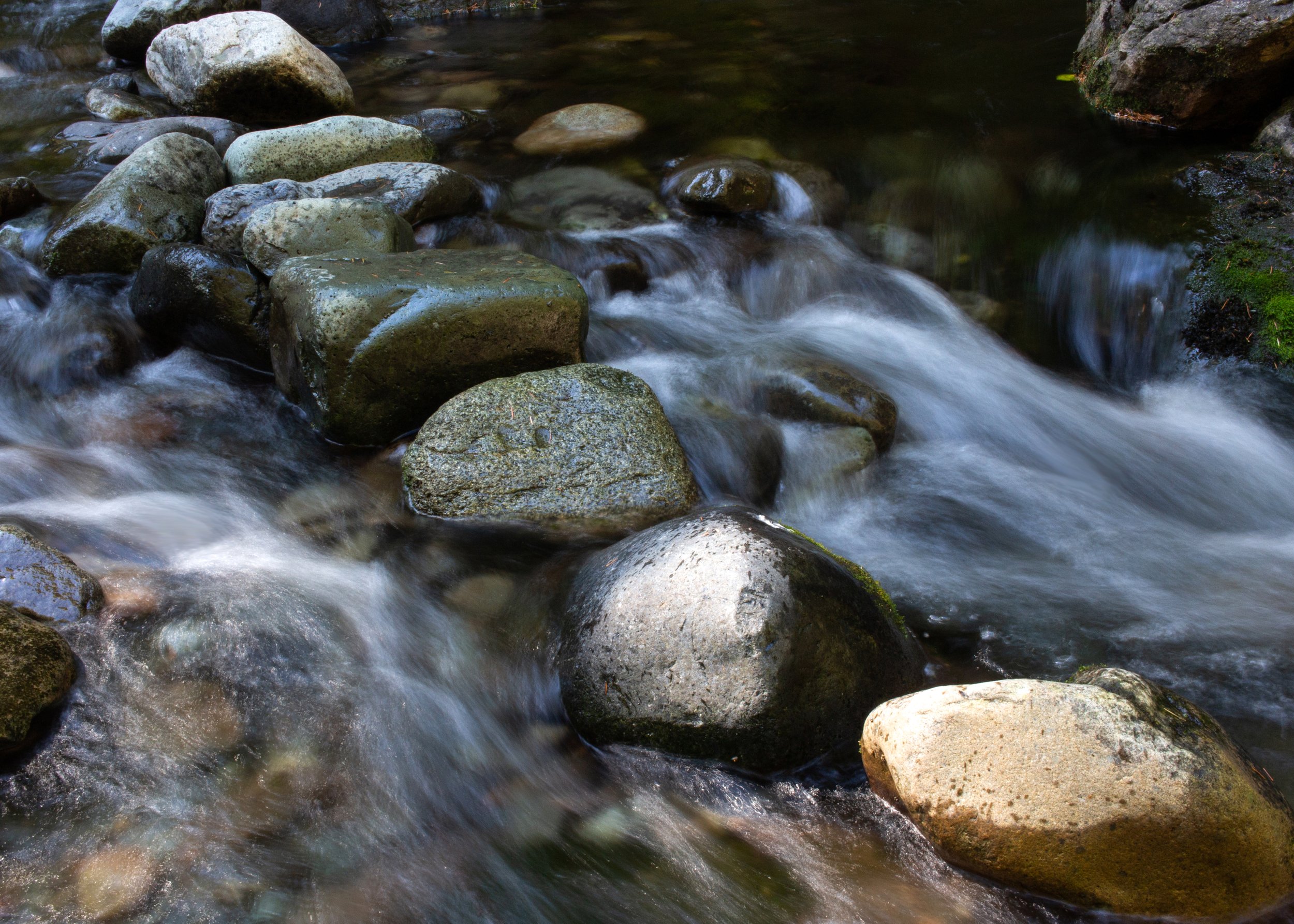 A stream with moving water flowing over and around various rounded rocks and stones.