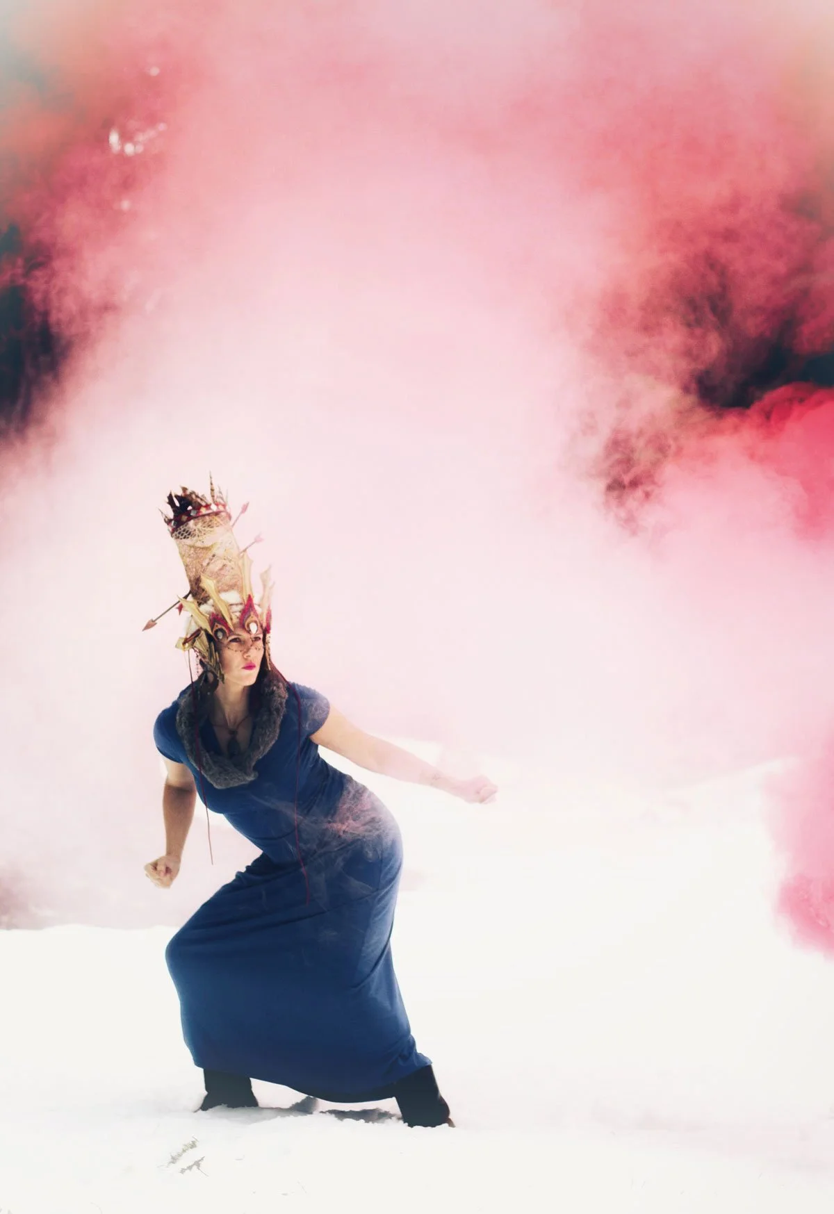 A woman standing in snow wearing a long blue dress and a tall, ornate headdress, with pink and black smoke in the background.