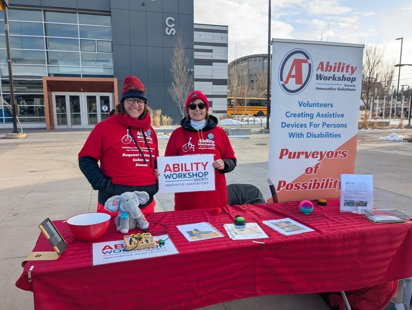 Two women wearing red hoodies and winter hats stand behind a table at an outdoor event, promoting the Ability Workshop Society for creating assistive devices for persons with disabilities. The table displays various items and informational materials, with a banner indicating the organization’s mission.