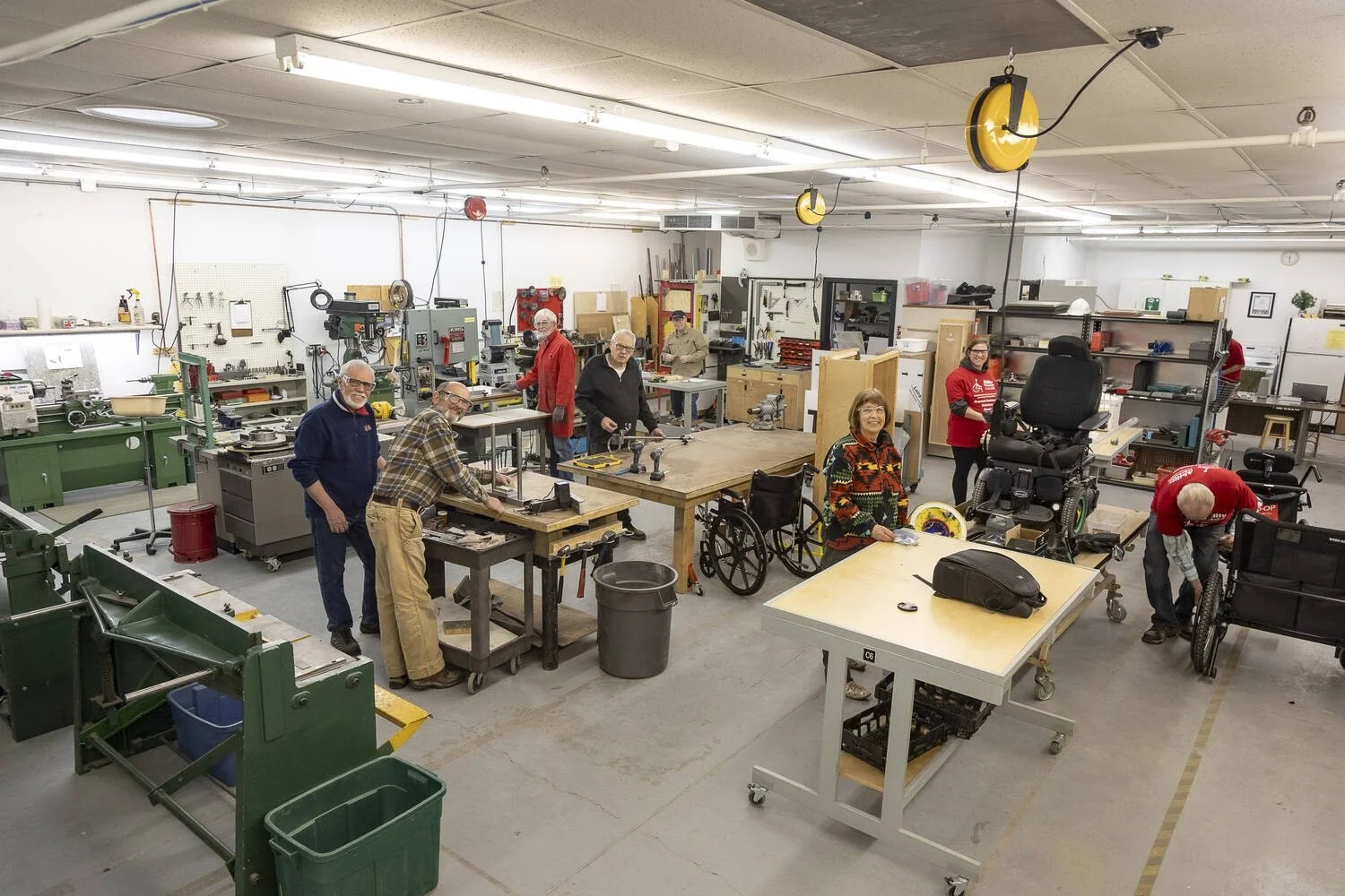 People working in a spacious workshop with various tools, machinery, and workbenches, some in wheelchairs, smiling at the camera.