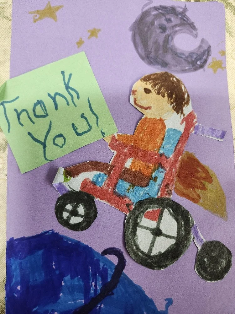 A colorful drawing of a person in a wheelchair with a green sticky note that says 'Thank You!' The background features a purple sky with a moon and stars, and blue ground with what seems to be a black animal tail or leg.