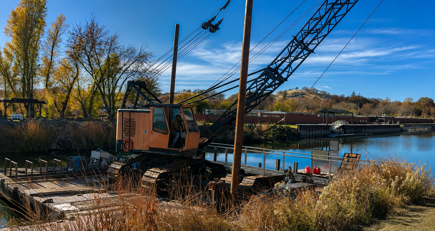A construction excavator with a long boom arm working near a body of water with trees, hills, and a clear blue sky in the background.
