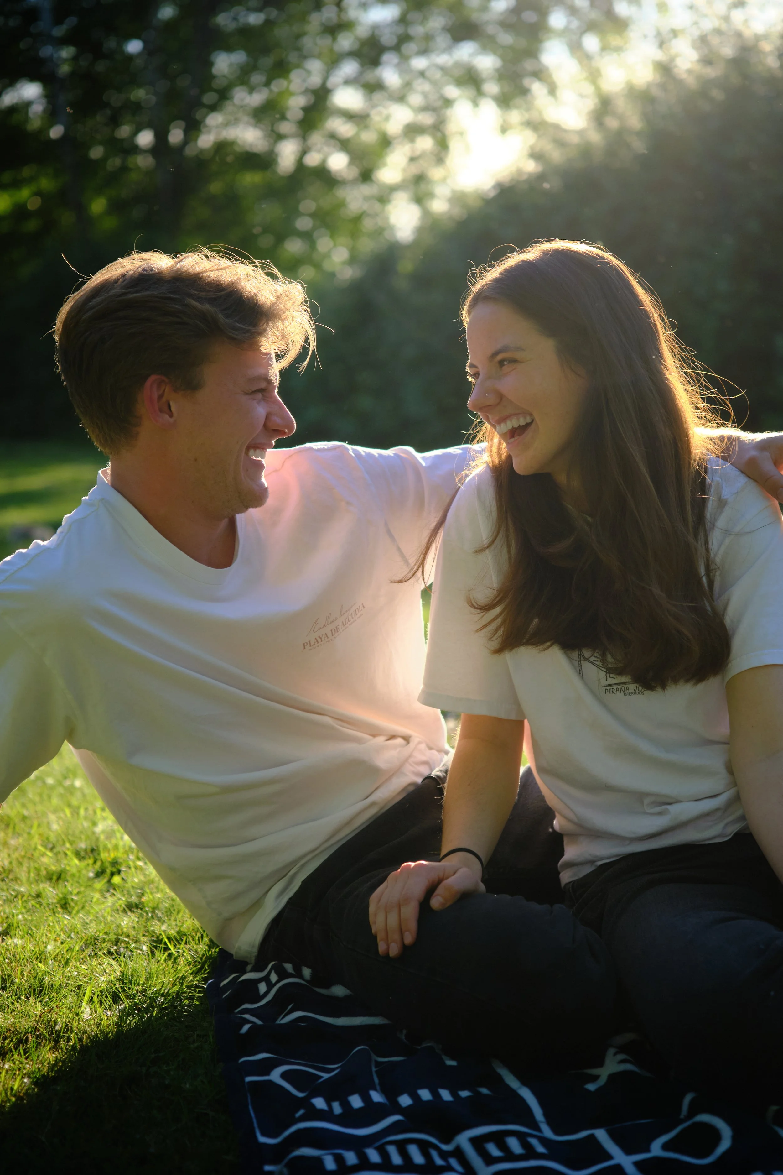 A young man and woman sitting on grass outdoors, smiling and looking at each other, with trees and sunlight in the background.