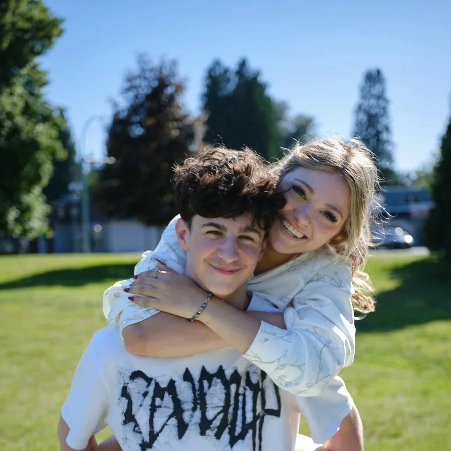 A young man giving a piggyback ride to a young woman in a park on a sunny day, smiling and hugging.
