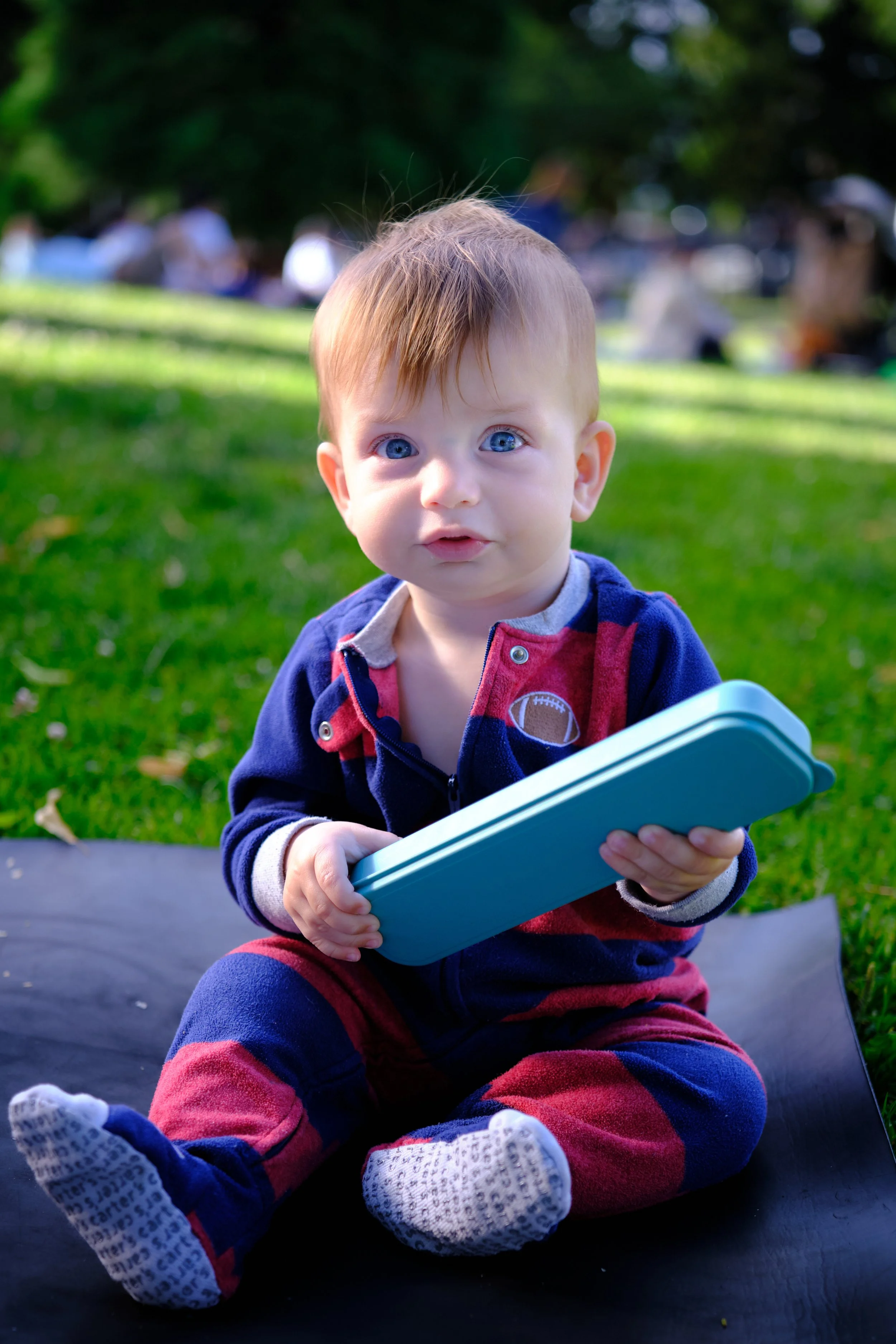 A young child with blue eyes, wearing a navy and red outfit, sitting outdoors on a black surface. The child is holding a blue tablet and looking at the camera with a curious expression. In the background, there are blurry trees and people, indicating