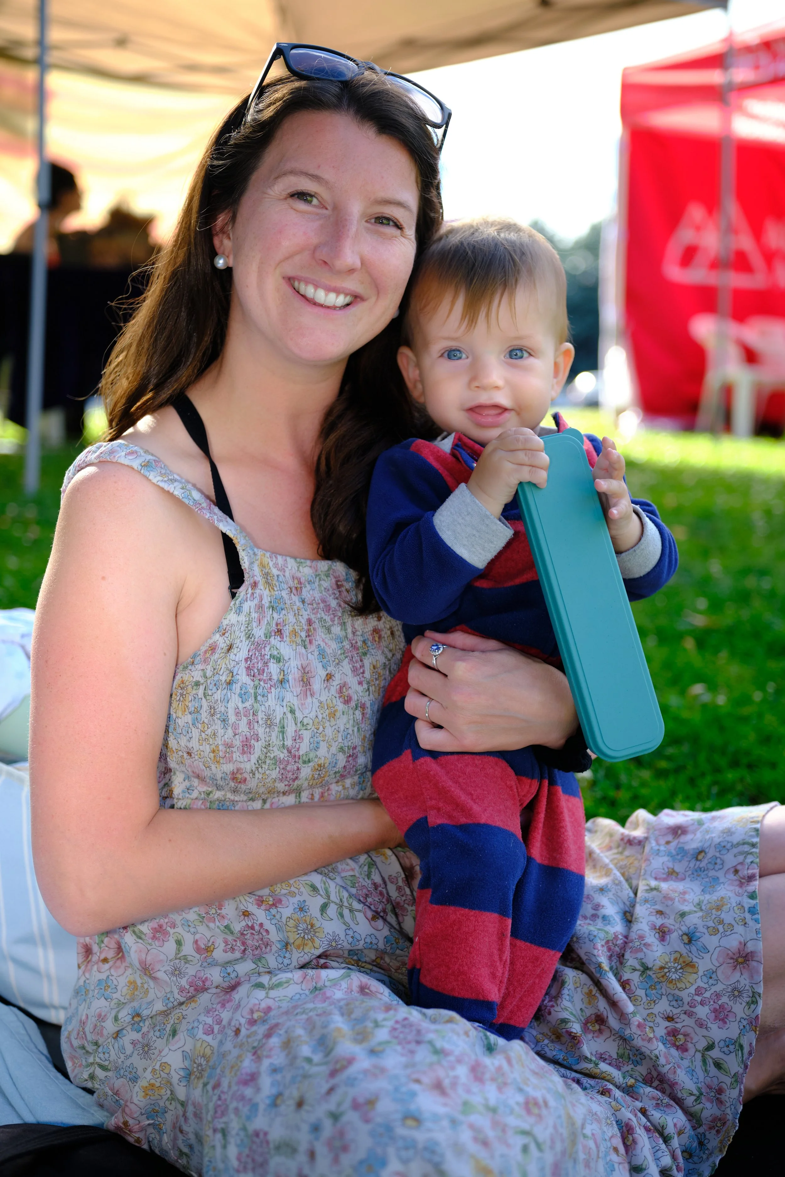 A smiling woman with long brown hair, wearing pearl earrings and sunglasses on her head, holding a young boy with blue eyes and light brown hair, dressed in a striped red and blue outfit, sitting outdoors on a sunny day with tents and a grassy area i