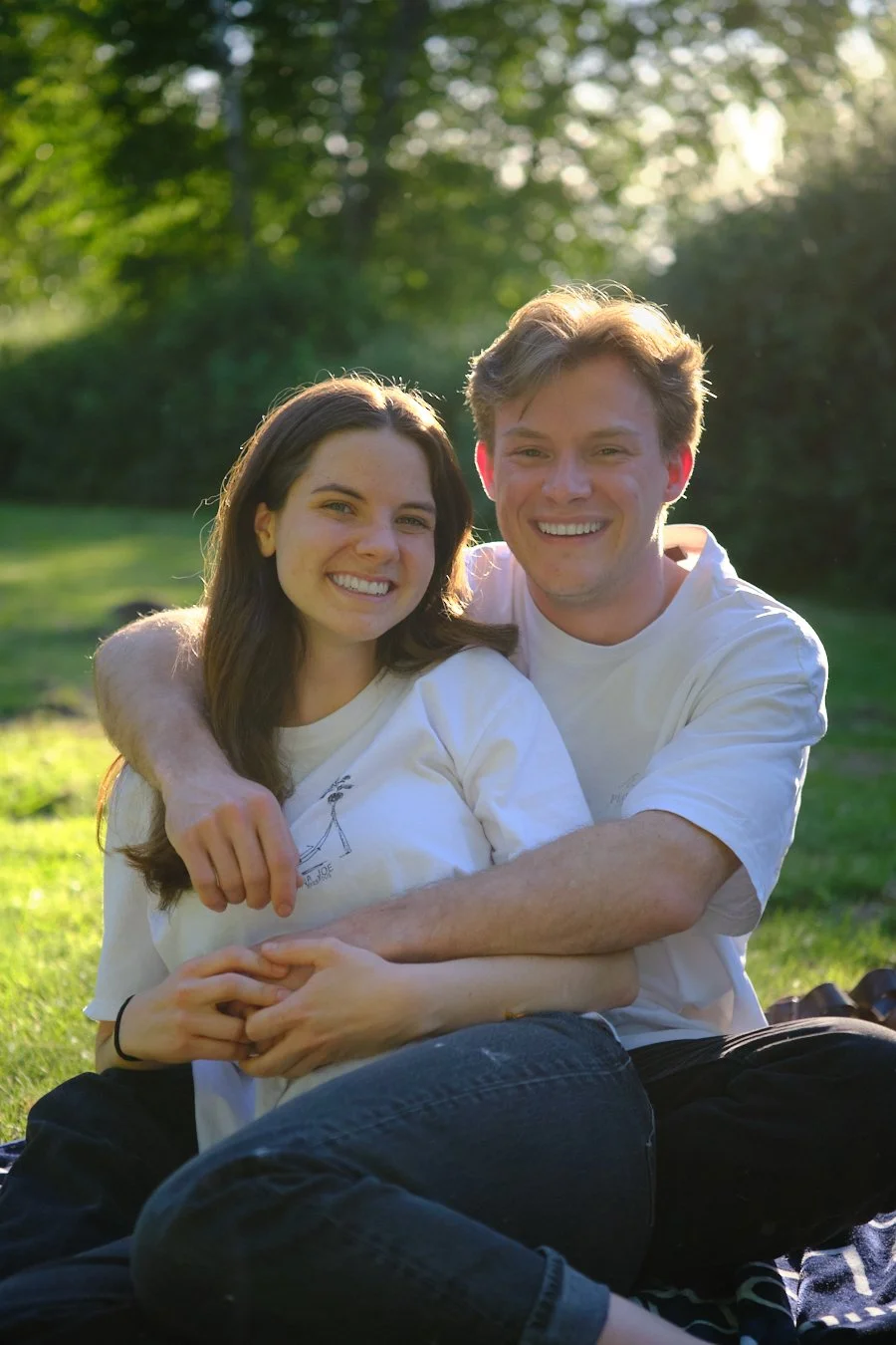 A smiling couple sitting outdoors on grass, embracing each other in the sunlight with trees in the background.