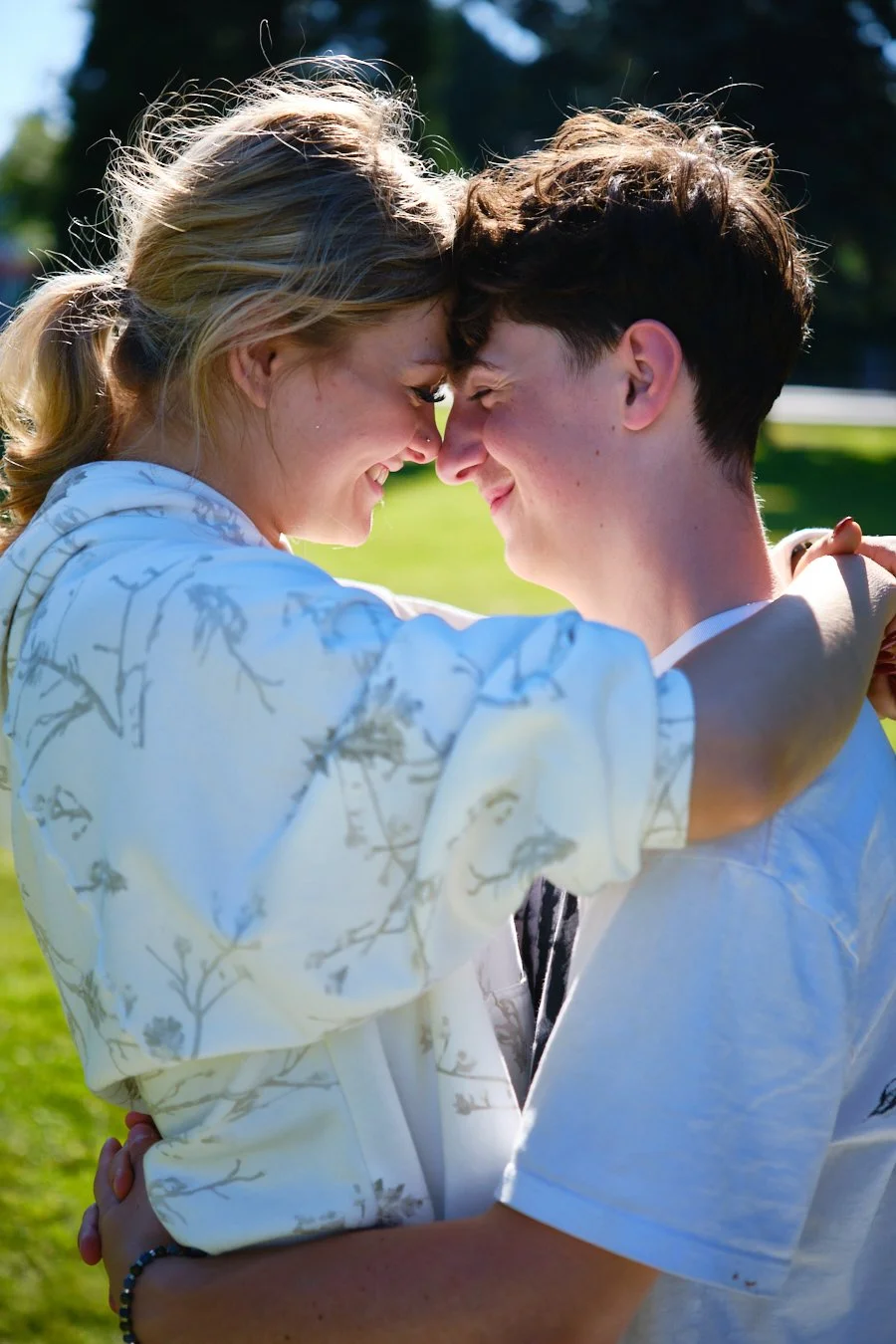 A young couple is smiling and touching foreheads outdoors on a sunny day, with green grass and trees in the background.