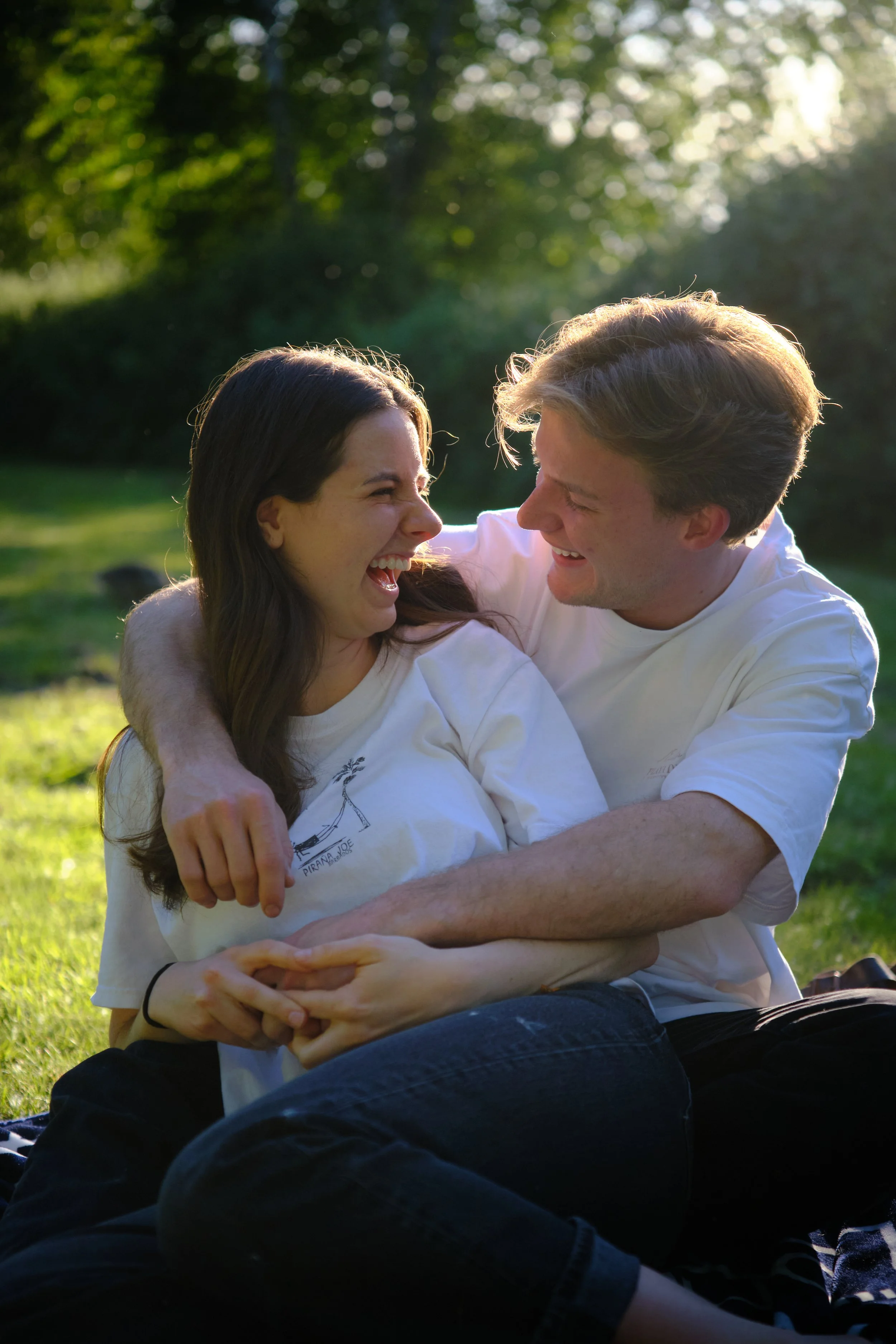 A young woman and a young man sit close together outdoors in a grassy area during sunset, smiling and laughing with their faces close, embracing each other.