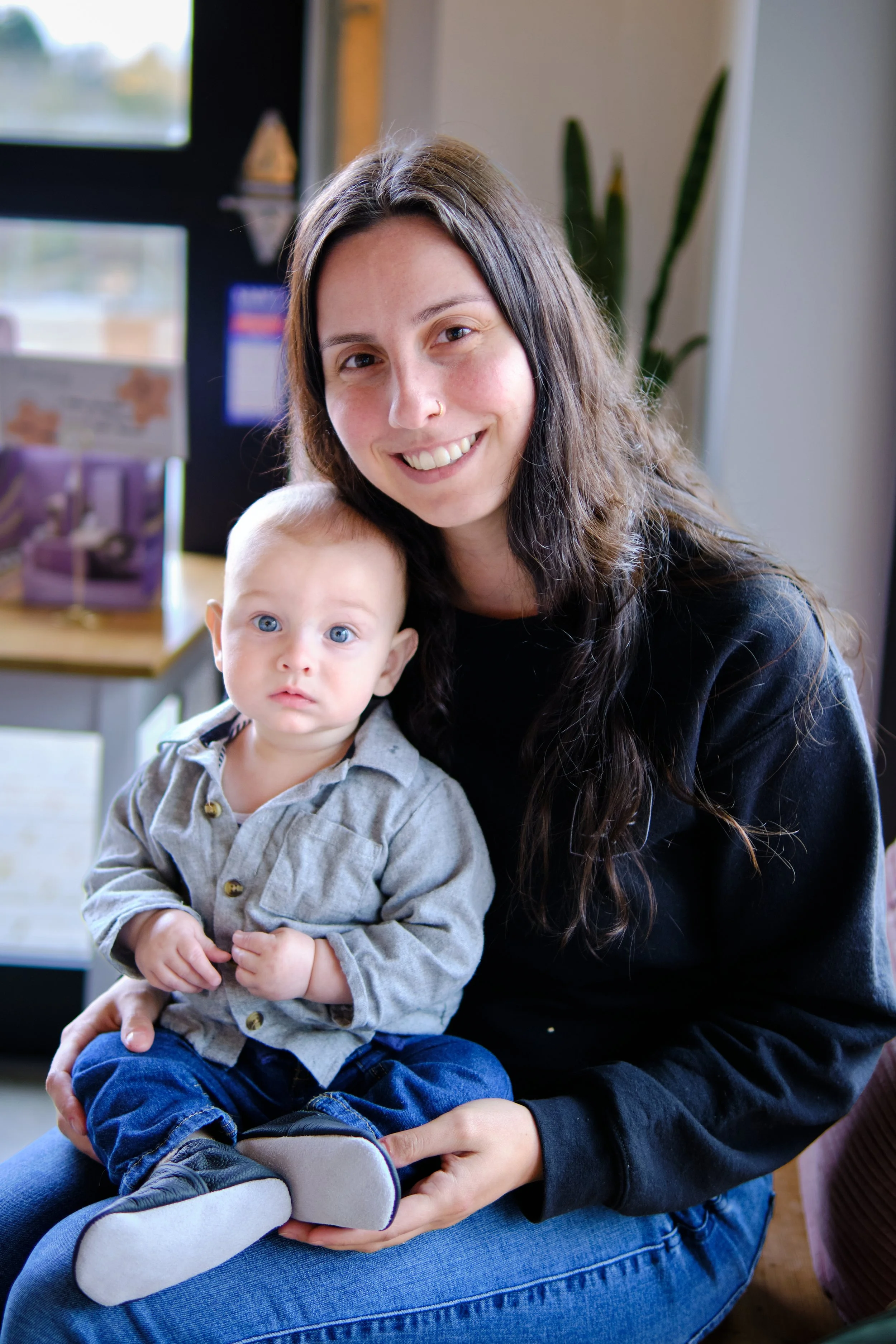 A woman with long brown hair, smiling, holding a young boy with blue eyes and light hair, both sitting indoors.