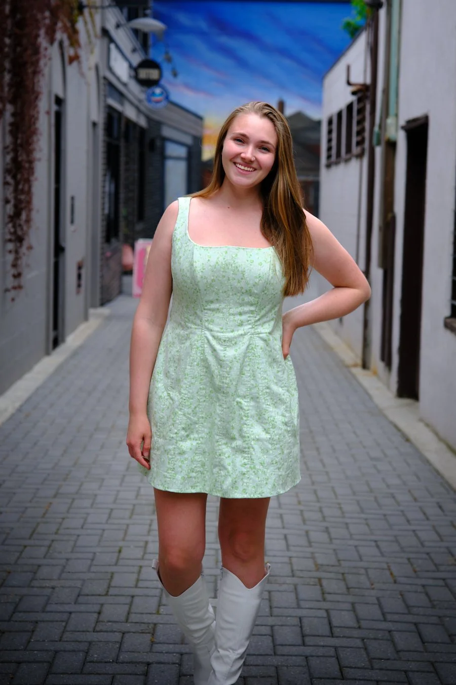 A young woman with long, blond hair wearing a green patterned sleeveless dress and white knee-high boots, smiling and standing in a narrow alleyway with buildings and a colorful mural in the background.