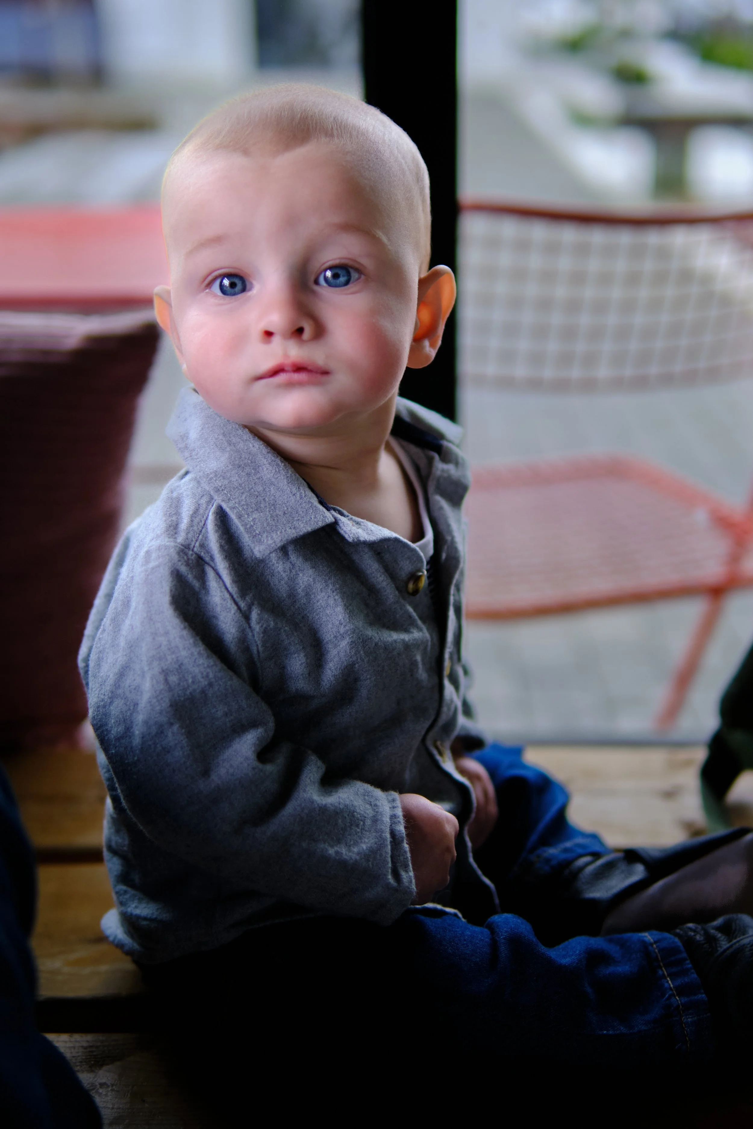 A young boy with bright blue eyes and a shaved head sitting indoors on a bench, looking towards the camera.